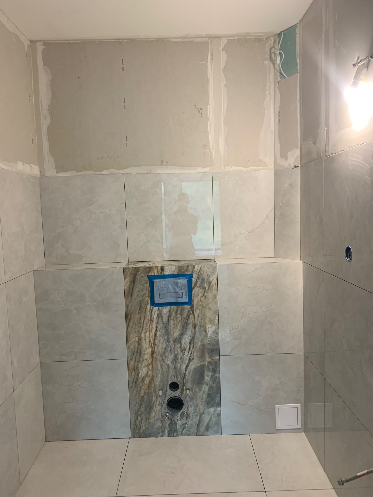 Płytki imitujące marmur na ścianie łazienki w trakcie remontu, widoczne otwory na armaturę i fragment instalacji podtynkowej WC.