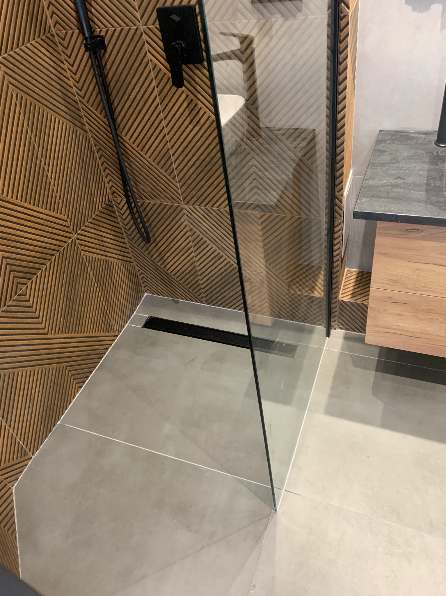 Nowoczesna kabina prysznicowa typu walk-in z odpływem liniowym, wykończona płytkami imitującymi drewno o geometrycznym wzorze i szarą posadzką.