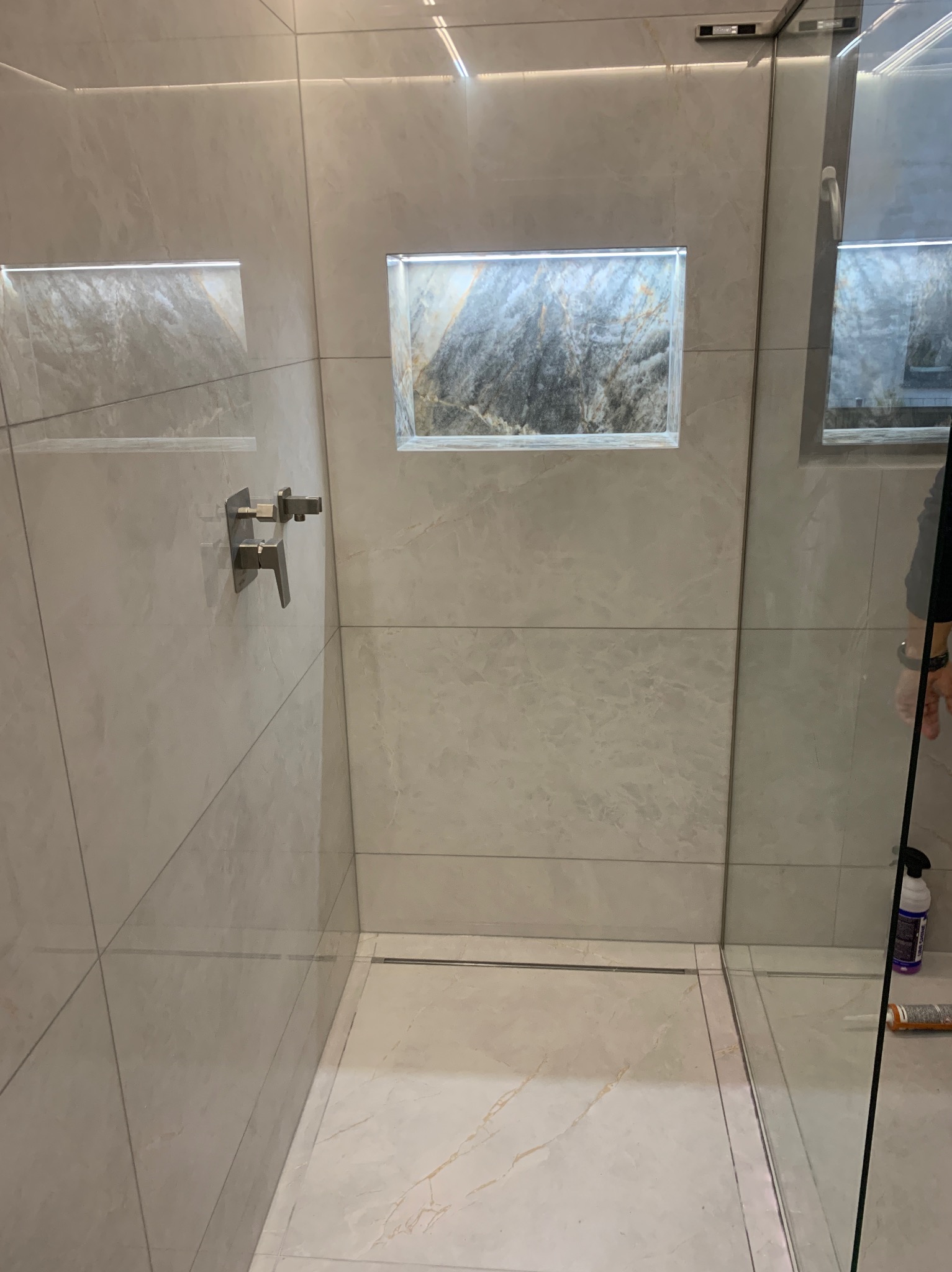 Nowoczesna kabina prysznicowa z odpływem liniowym, wykończona płytkami imitującymi marmur, z wnękami podświetlanymi LED i armaturą w kolorze stali szczotkowanej.