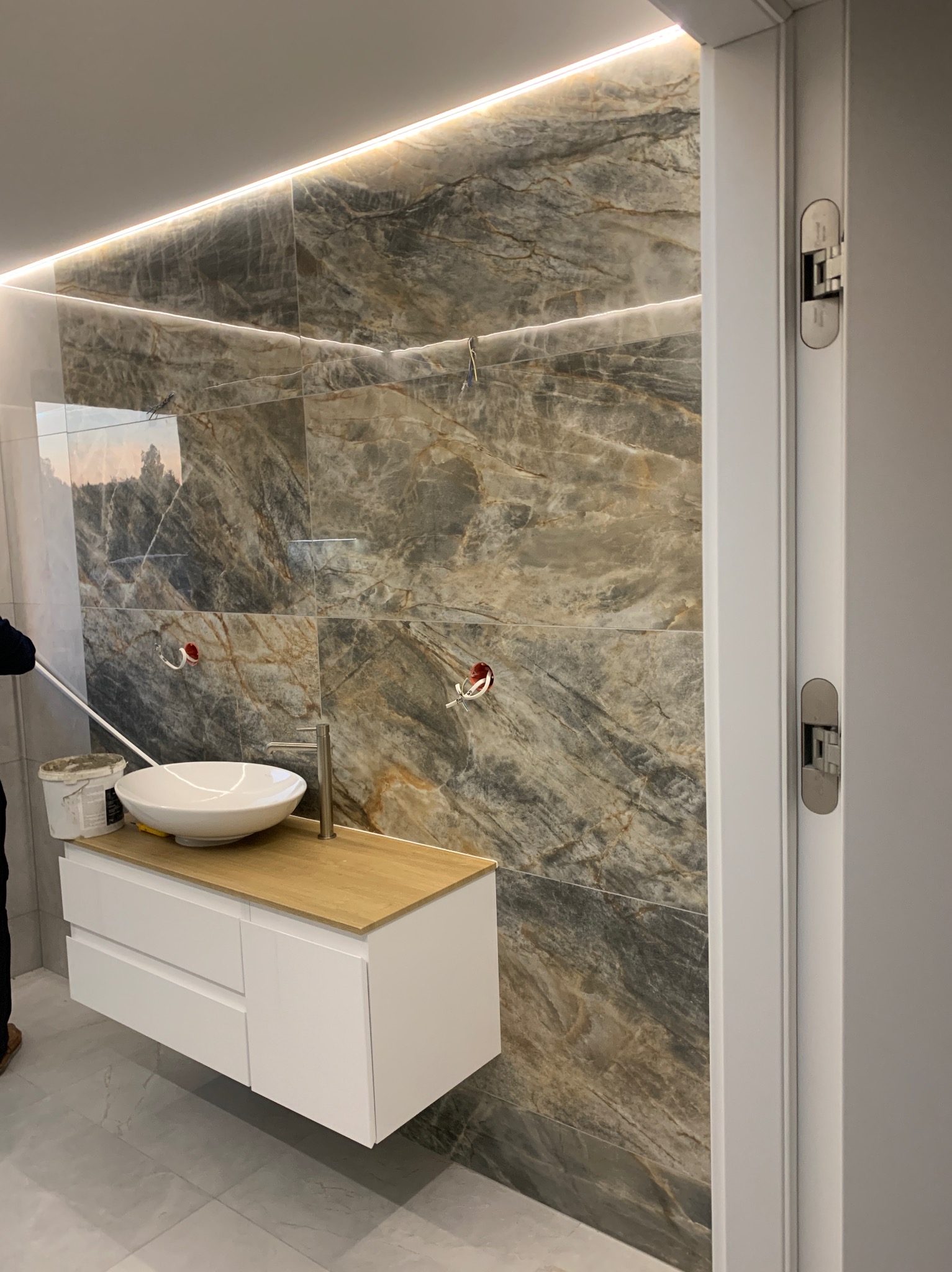 Nowoczesna łazienka w trakcie wykończenia: umywalka nablatowa na białej szafce, marmurowe płytki na ścianie z oświetleniem LED i widoczne elementy instalacji hydraulicznej.