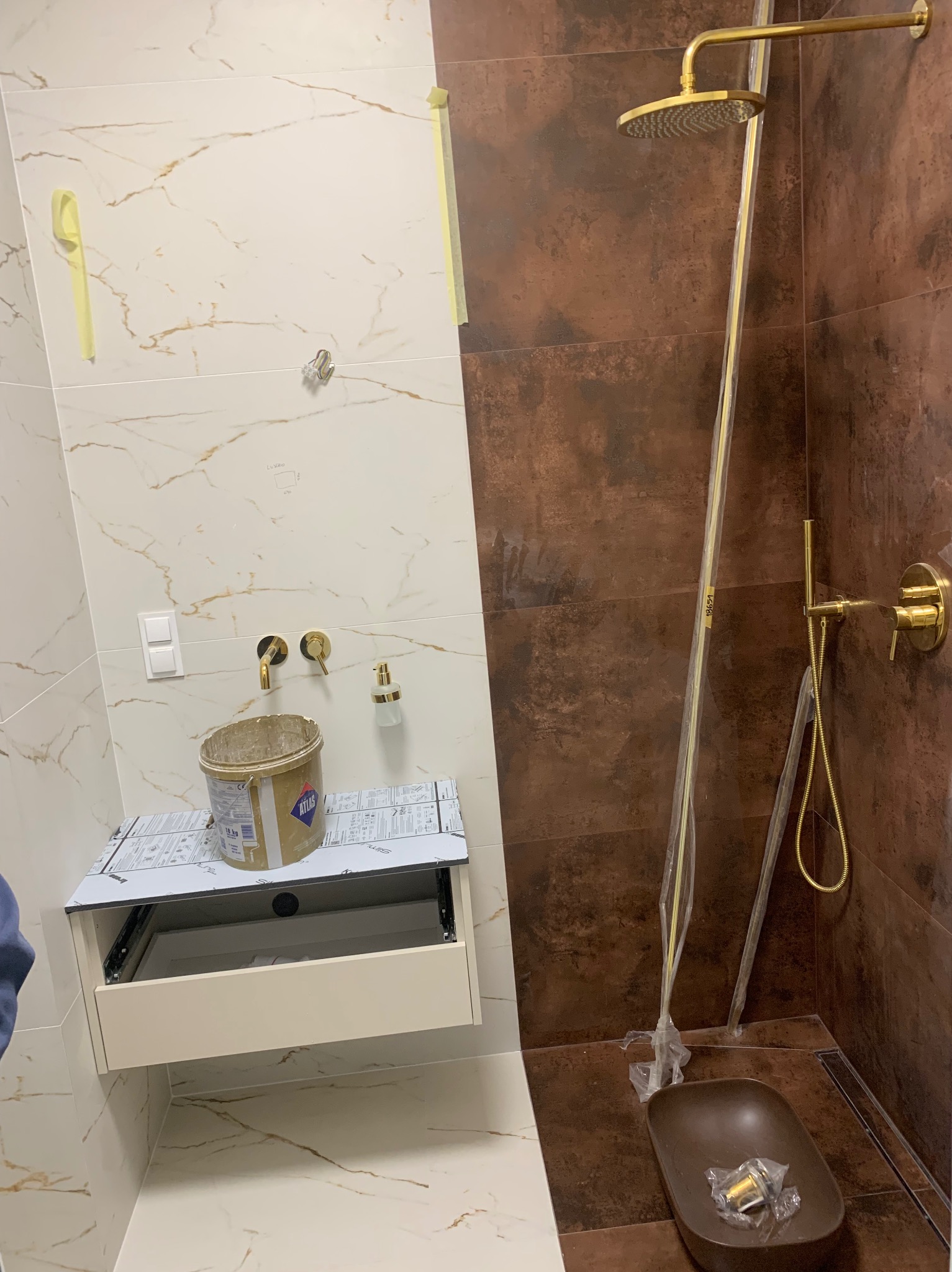 Montaż złotej armatury w łazience z brązowymi i białymi płytkami, widoczna umywalka na szafce z otwartą szufladą, wiadro po kleju do płytek oraz elementy kabiny prysznicowej w folii ochronnej.