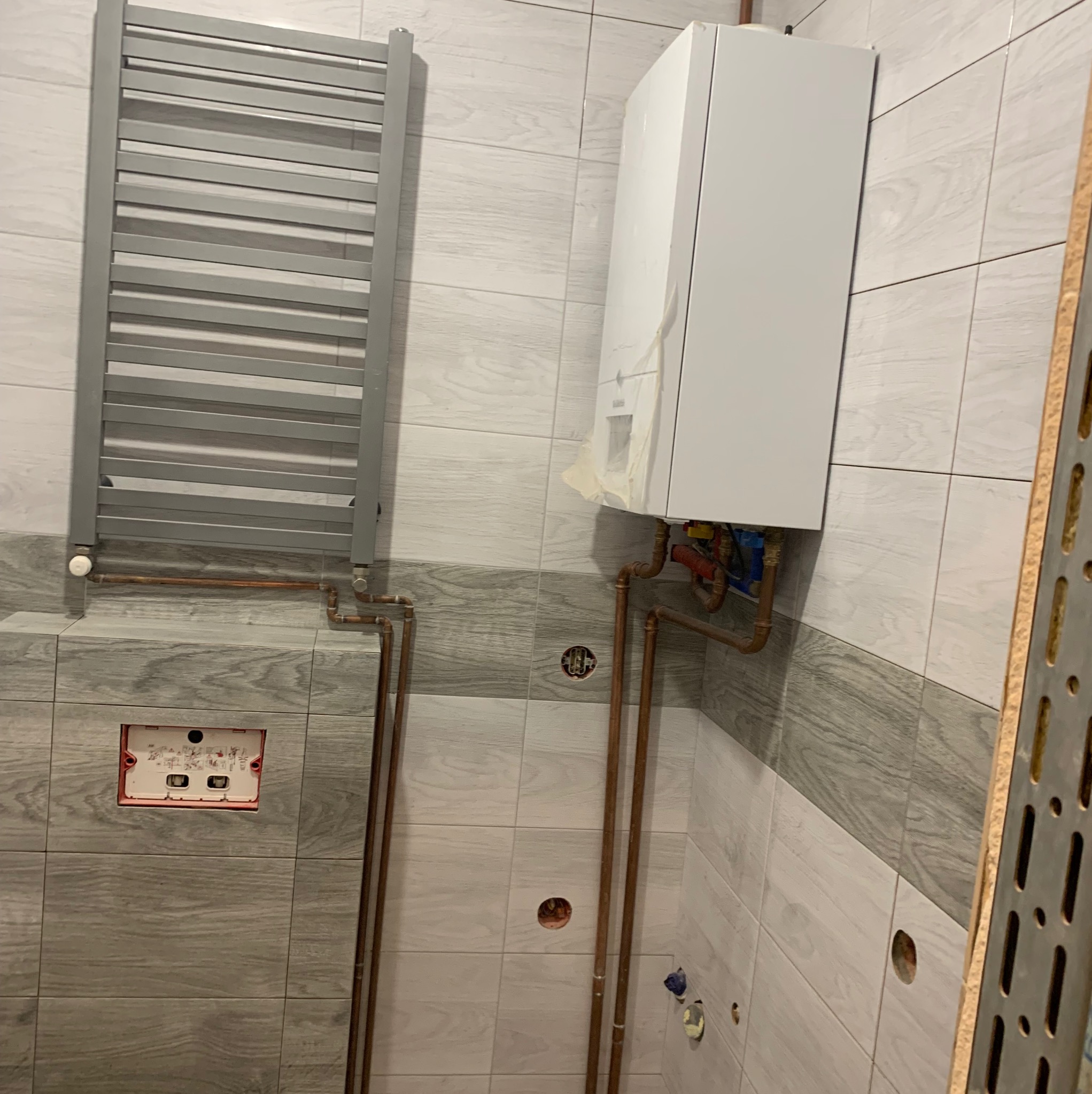 Biały piec gazowy kondensacyjny, szary grzejnik drabinkowy i miedziane rury w trakcie montażu w wykończonej łazience z szaro-białymi płytkami imitującymi drewno.