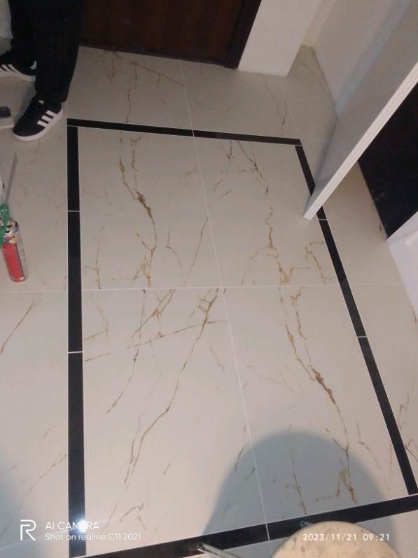 Ułożone płytki podłogowe w łazience, kremowe z brązowym marmurkowym wzorem, obramowane czarnymi paskami, widoczne fragmenty butów i puszka z pianką montażową.