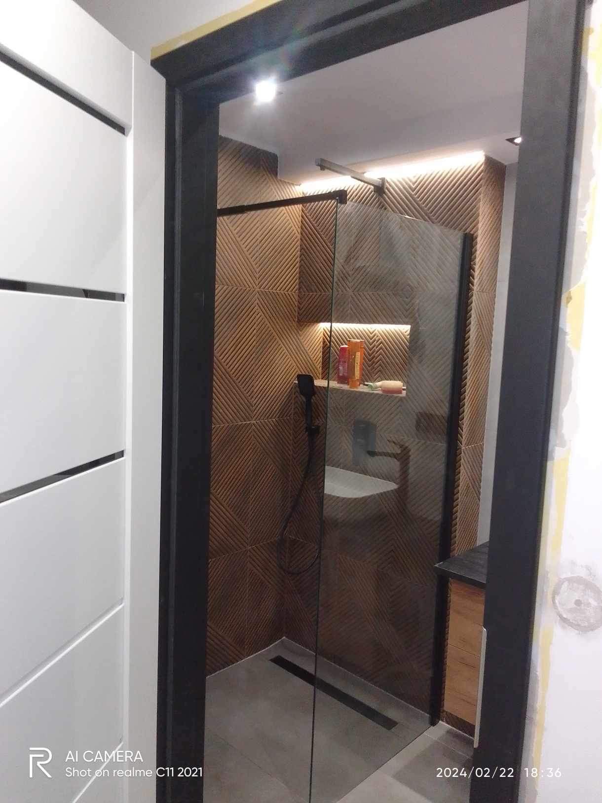 Nowoczesna kabina prysznicowa walk-in z czarną armaturą i odpływem liniowym, brązowe płytki z geometrycznym wzorem, podświetlana półka z kosmetykami, widok z zewnątrz kabiny.