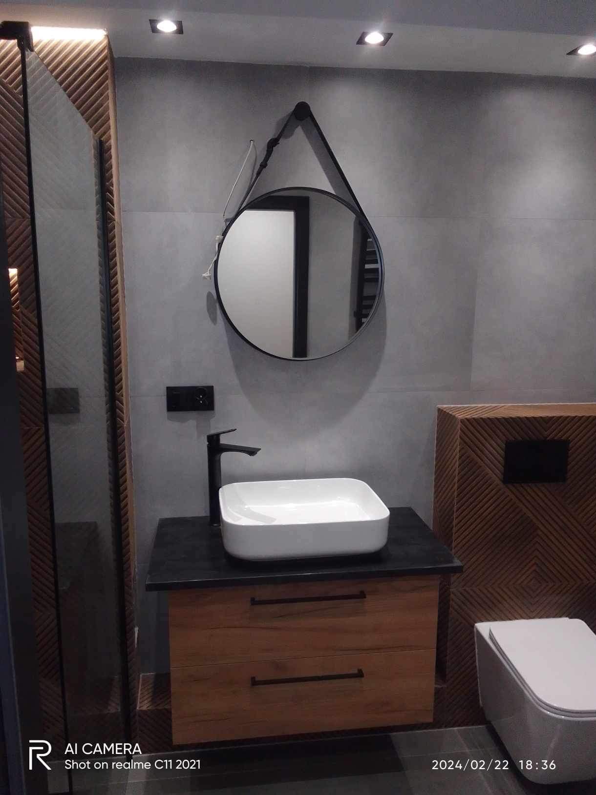 Nowoczesna łazienka z umywalką nablatową, czarną armaturą i okrągłym lustrem na pasku, drewniana szafka pod umywalką z szufladami, szara ściana z płytkami i oświetleniem punktowym.