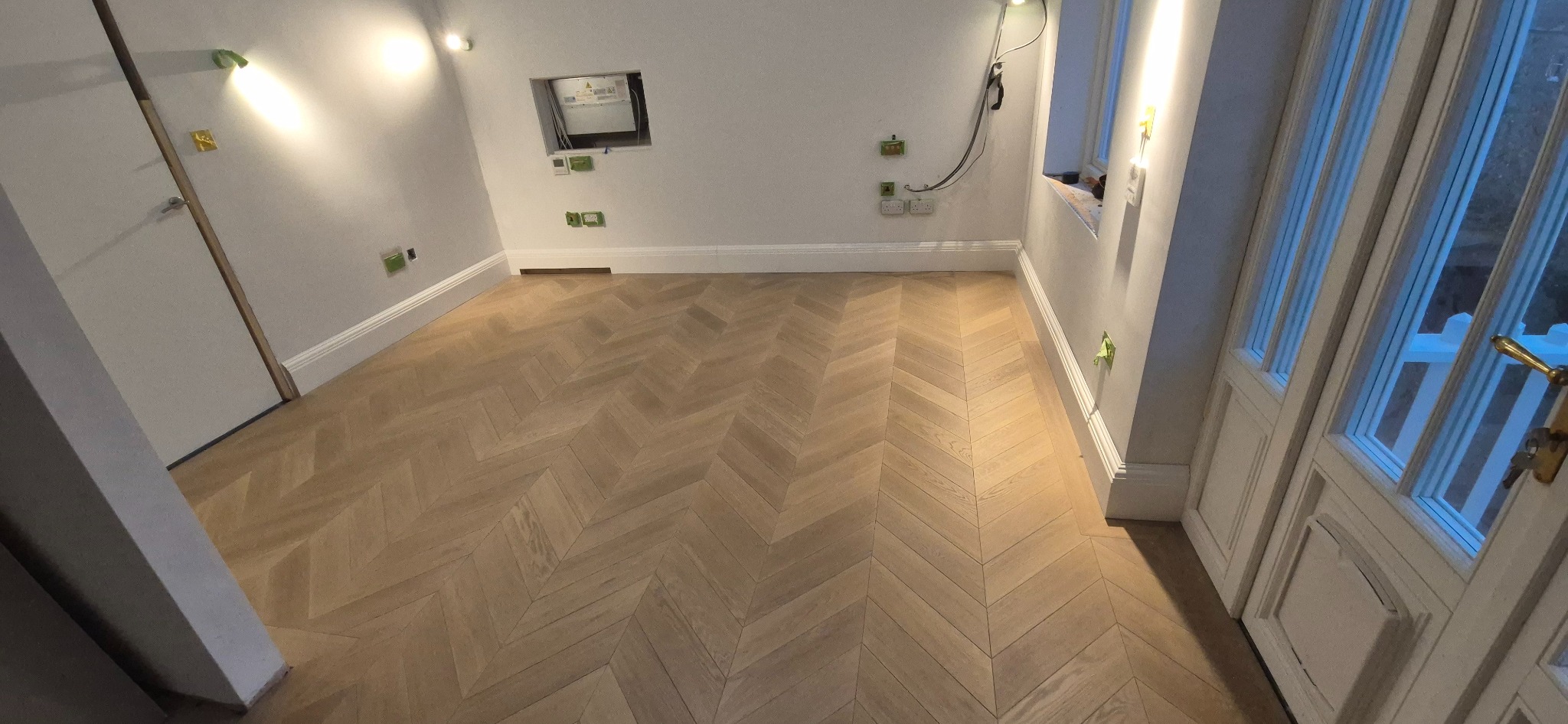 Jasny, nowo ułożony parkiet w jodełkę w pustym pokoju z białymi ścianami, widoczne gniazdka elektryczne i otwór w ścianie z przewodami.