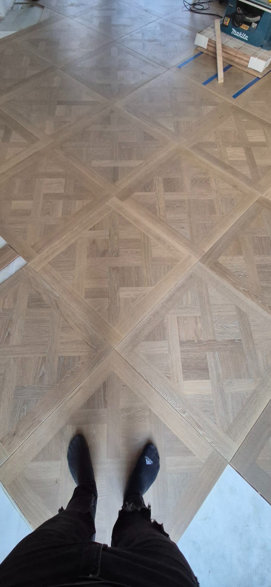 Nowa, jasna podłoga parkietowa ułożona we wzór kwadratów, widok z góry na stopy w czarnych skarpetkach i fragment spodni, w tle maszyna do cięcia drewna i listwy przypodłogowe.