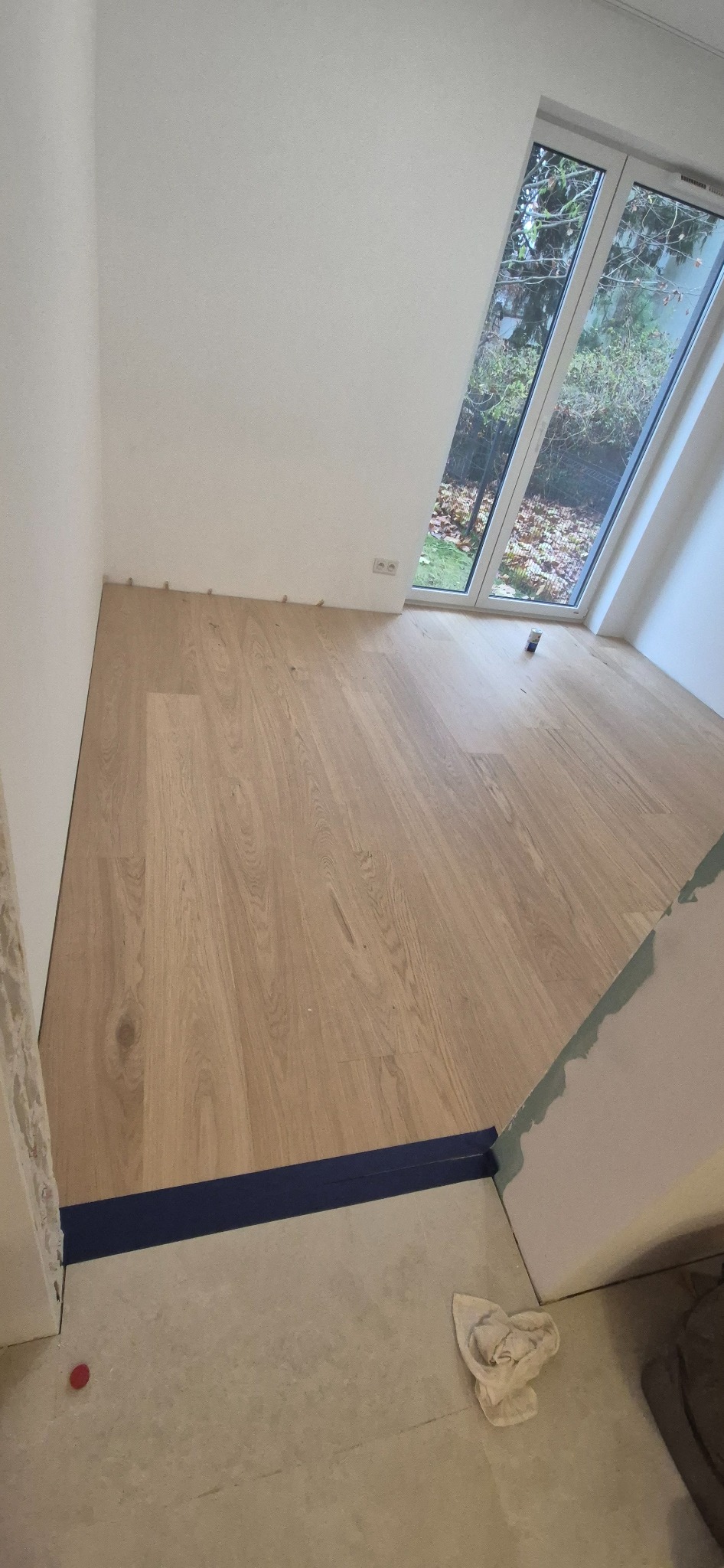 Fragment pokoju z jasną, drewnianą podłogą w trakcie montażu, widoczne listwy przypodłogowe, okno dachowe z widokiem na zieleń oraz drobne elementy wykończeniowe.