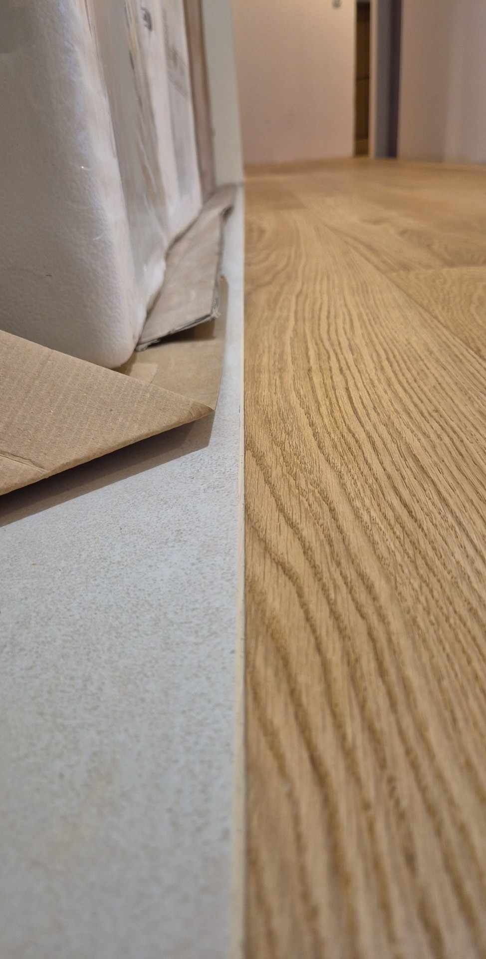 Fragment podłogi z jasnego drewna sąsiadujący z zabezpieczoną folią ścianą i kartonem ochronnym przy listwie przypodłogowej, widoczne usłojenie drewna.