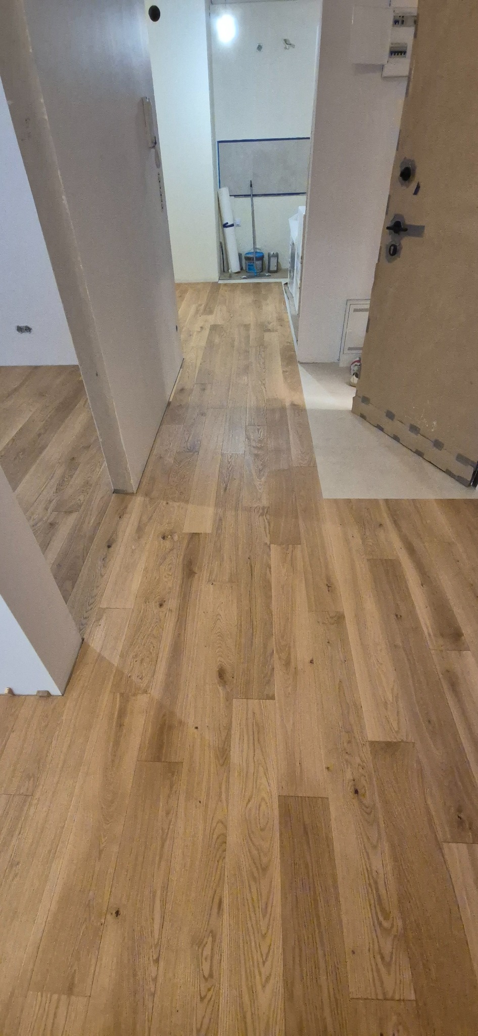 Świeżo ułożony parkiet w odcieniach naturalnego drewna, widoczny fragment progu drzwi i przygotowania do dalszych prac w tle.