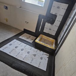 Wiktoria Iwańska Hydro-PUR Solution - Hydroizolacja w narożniku łazienki z widocznym odpływem liniowym, uszczelniona czarną folią w płynie i wzmocniona taśmami uszczelniającymi marki Ardex SK 100 W.