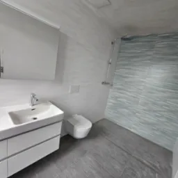 Nowoczesna łazienka w odcieniach szarości i bieli, z umywalką na szafce, toaletą i prysznicem z deszczownicą, wykończona płytkami imitującymi kamień i drewno.