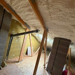 Poddasze po ociepleniu pianką PUR: widoczne drewniane krokwie, izolacja piankowa na skosach dachu, fragment instalacji wentylacyjnej i wełny mineralnej na ścianie szczytowej.