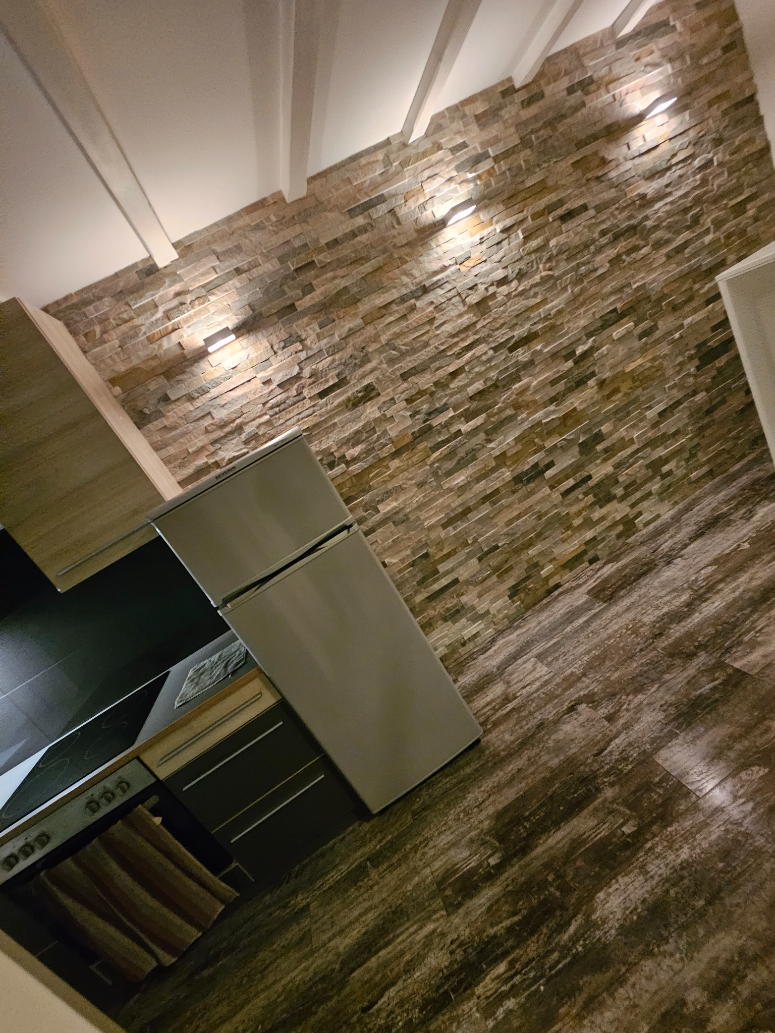 Kuchnia z kamienną ścianą, oświetleniem punktowym, drewnianą podłogą i srebrną lodówką Severin.