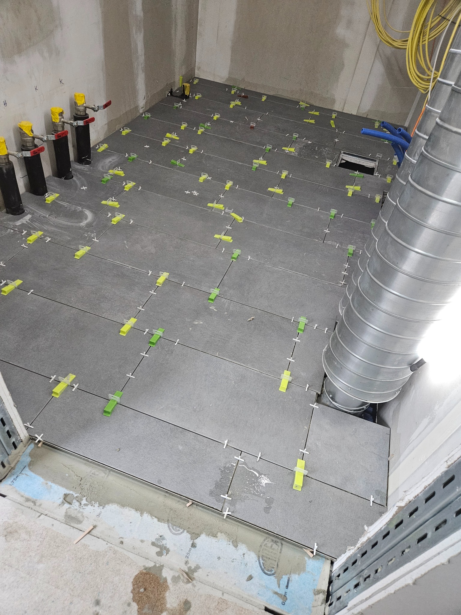 Szare płytki podłogowe w trakcie układania, z użyciem żółto-zielonych klinów poziomujących, widoczne pionowe rury z zaworami i przewody wentylacyjne.