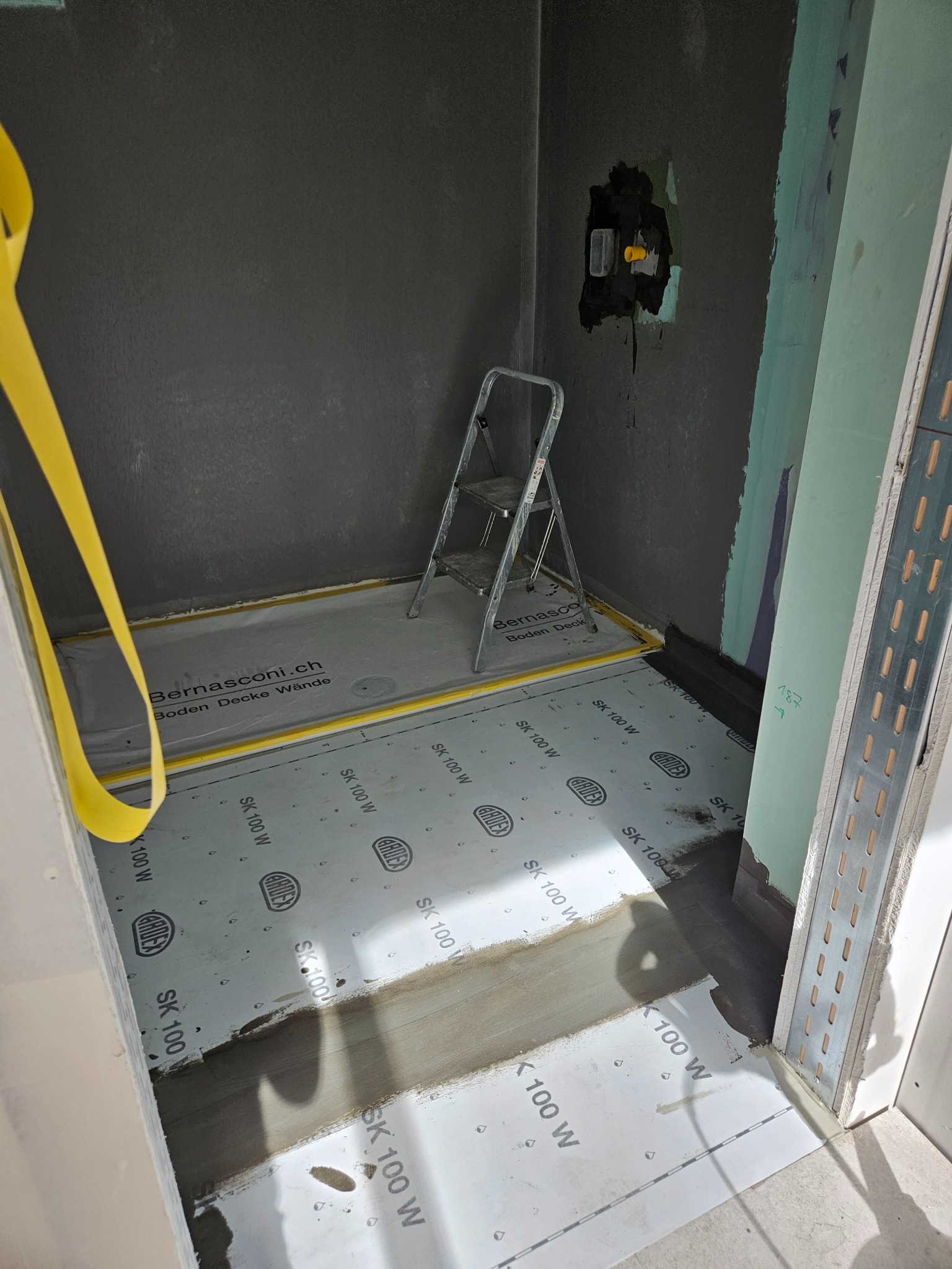 Przygotowanie podłoża w łazience: folia w płynie Ardex SK 100 W na podłodze, zabezpieczenie taśmą żółtą, ściany w surowym tynku, drabina aluminiowa w rogu, instalacja elektryczna przygotowana.