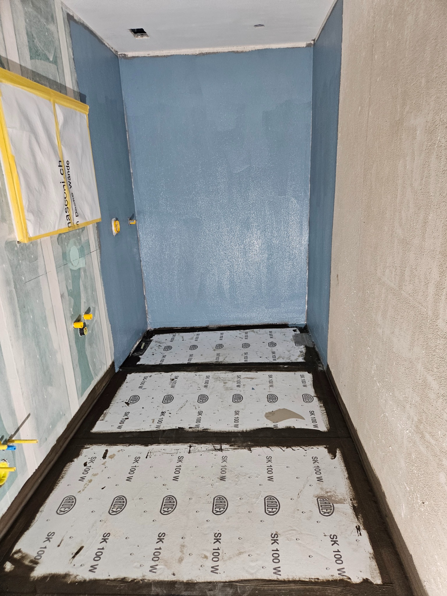 Przygotowanie do hydroizolacji w łazience: ściany pomalowane na niebiesko, na podłodze płyty hydroizolacyjne Ardex SK 100 W z widocznymi łączeniami, zabezpieczone narożniki. Na ścianie widoczne...