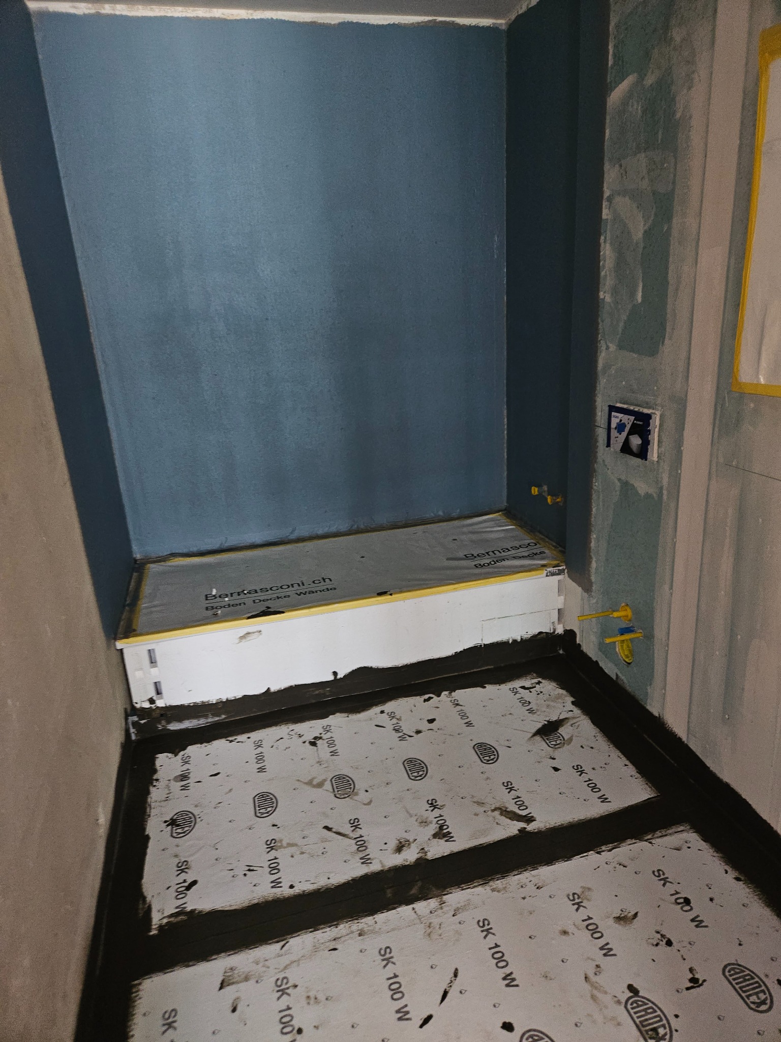 Przygotowanie podłoża pod płytki w łazience: hydroizolacja z użyciem maty uszczelniającej Ardex SK 100 W i taśmy uszczelniającej na styku ściany i podłogi, widoczne instalacje wodne i fragment...