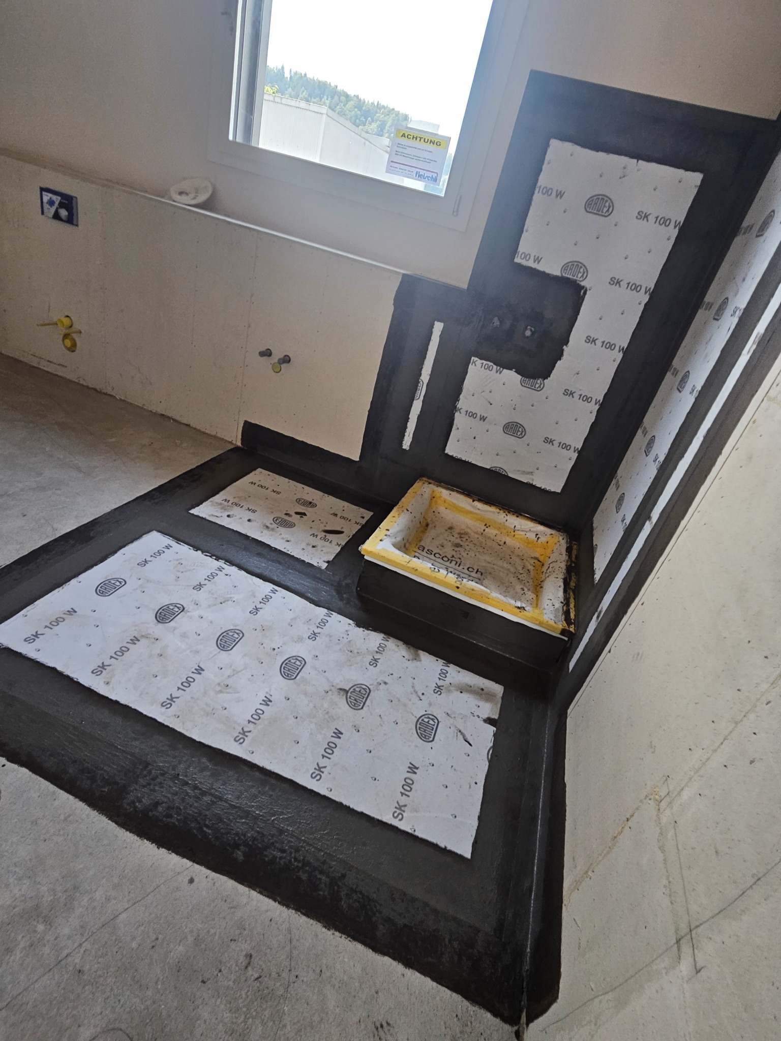 Hydroizolacja w narożniku łazienki z widocznym odpływem liniowym, uszczelniona czarną folią w płynie i wzmocniona taśmami uszczelniającymi marki Ardex SK 100 W.