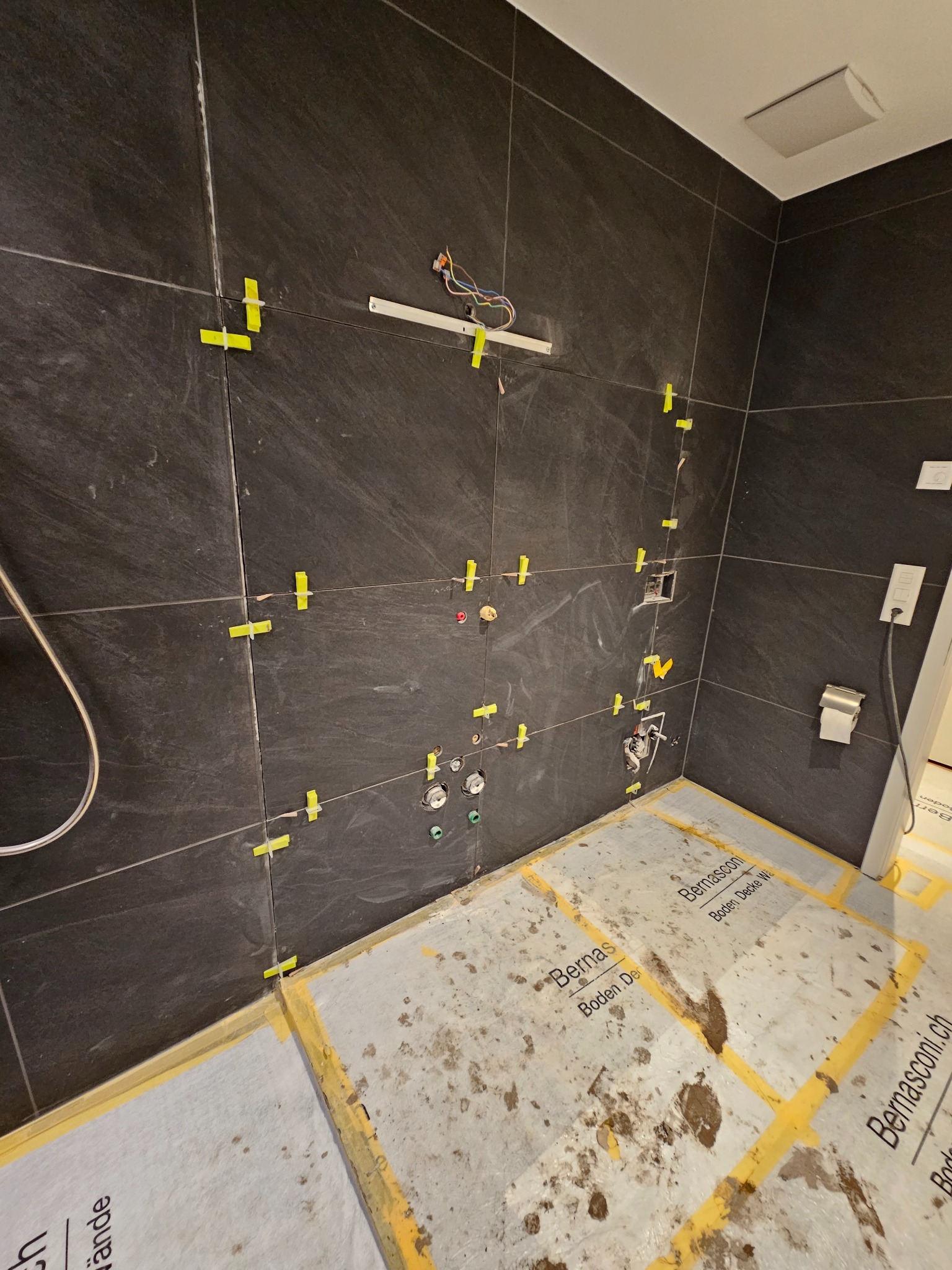 Łazienka w trakcie remontu: ciemne płytki imitujące kamień na ścianach, z żółtymi krzyżykami dystansowymi i taśmą malarską, zabezpieczona podłoga z widocznymi zabrudzeniami.
