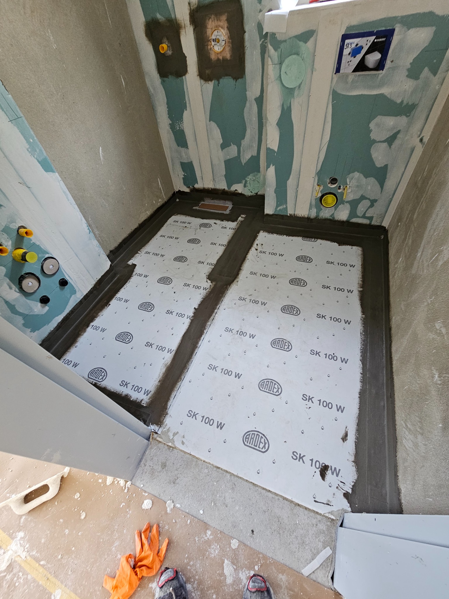 Przygotowanie podłoża w łazience: hydroizolacja z użyciem maty Ardex SK 100 W, widoczne rury instalacyjne w ścianie, pomarańczowa rękawica i buty robocze w dolnym rogu kadru.