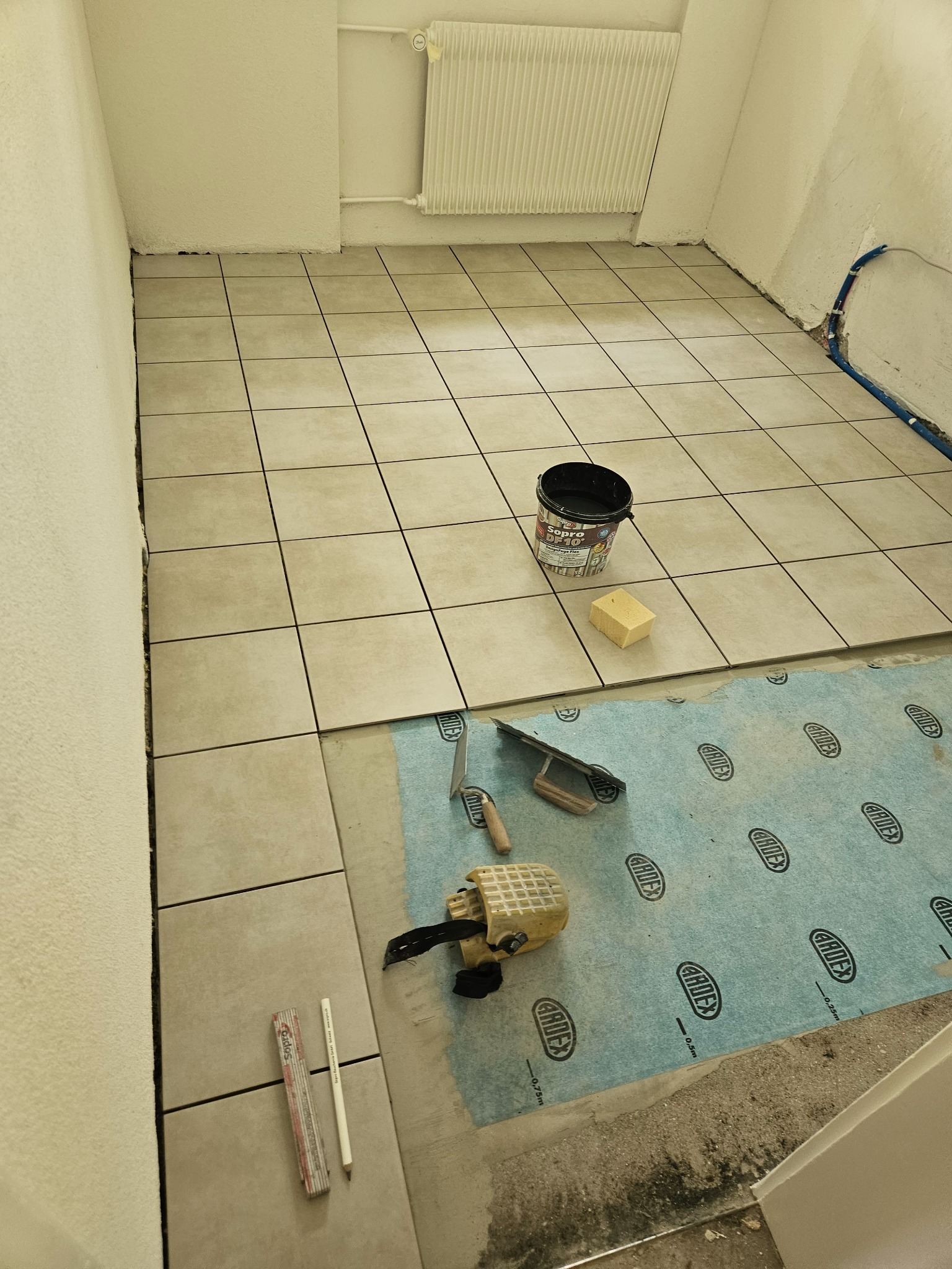 Wnętrze w trakcie układania beżowych płytek podłogowych, widoczne narzędzia: paca, wiadro z klejem Sopro DF 10, gąbka, miarka składana, ołówek, oraz niebieska mata pod płytki Ardex.