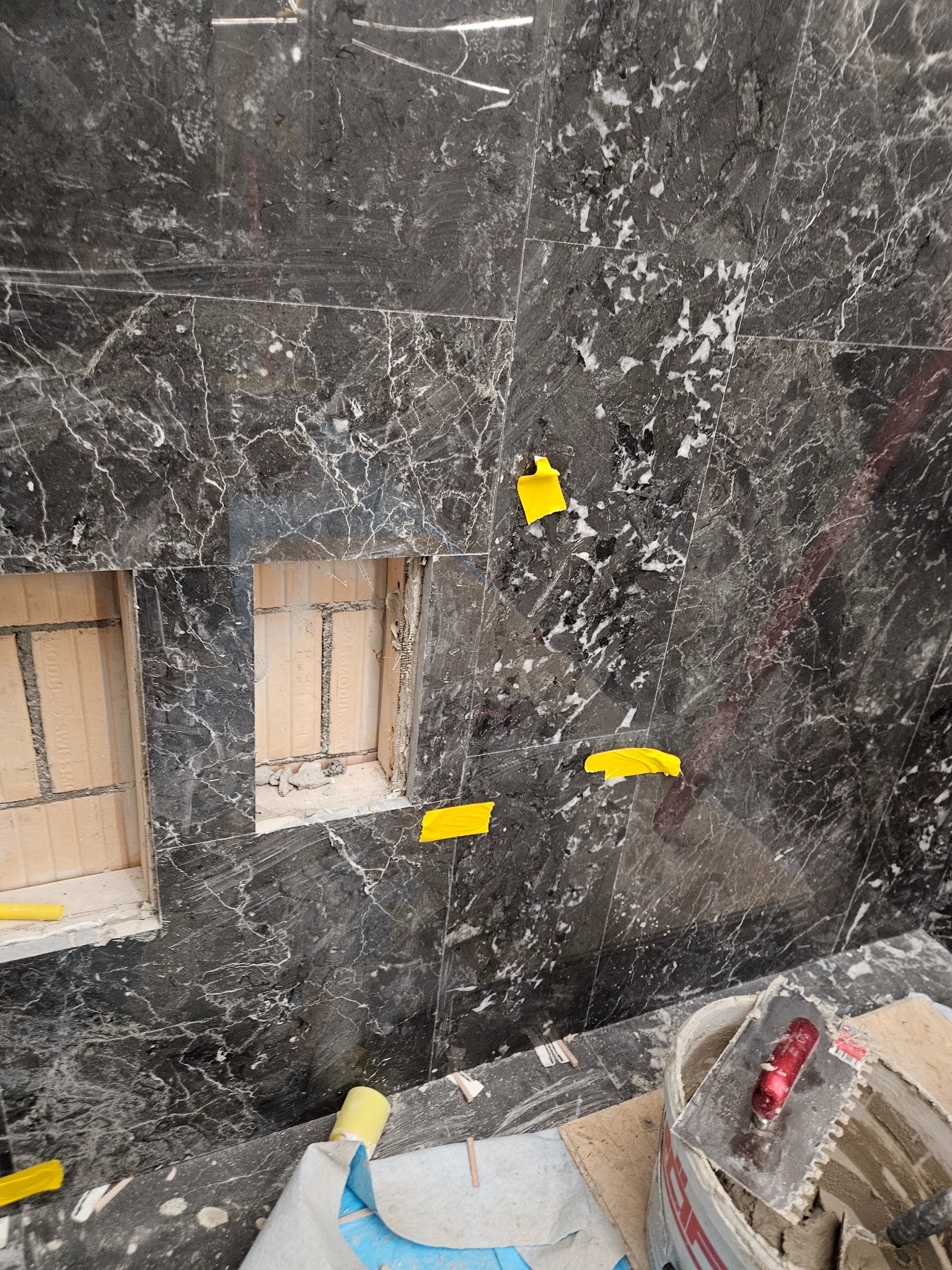 Montaż płytek imitujących czarny marmur z białymi żyłami, z wyciętymi wnękami, zabezpieczone żółtą taśmą malarską, widoczne narzędzia i zaprawa w wiadrze.