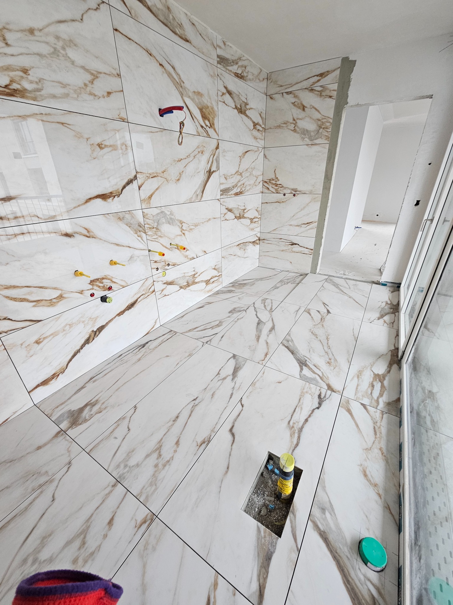 Wnętrze w trakcie prac wykończeniowych: marmurowe płytki na ścianach i podłodze, widoczne otwory na instalacje, fragment skarpety w rogu kadru.