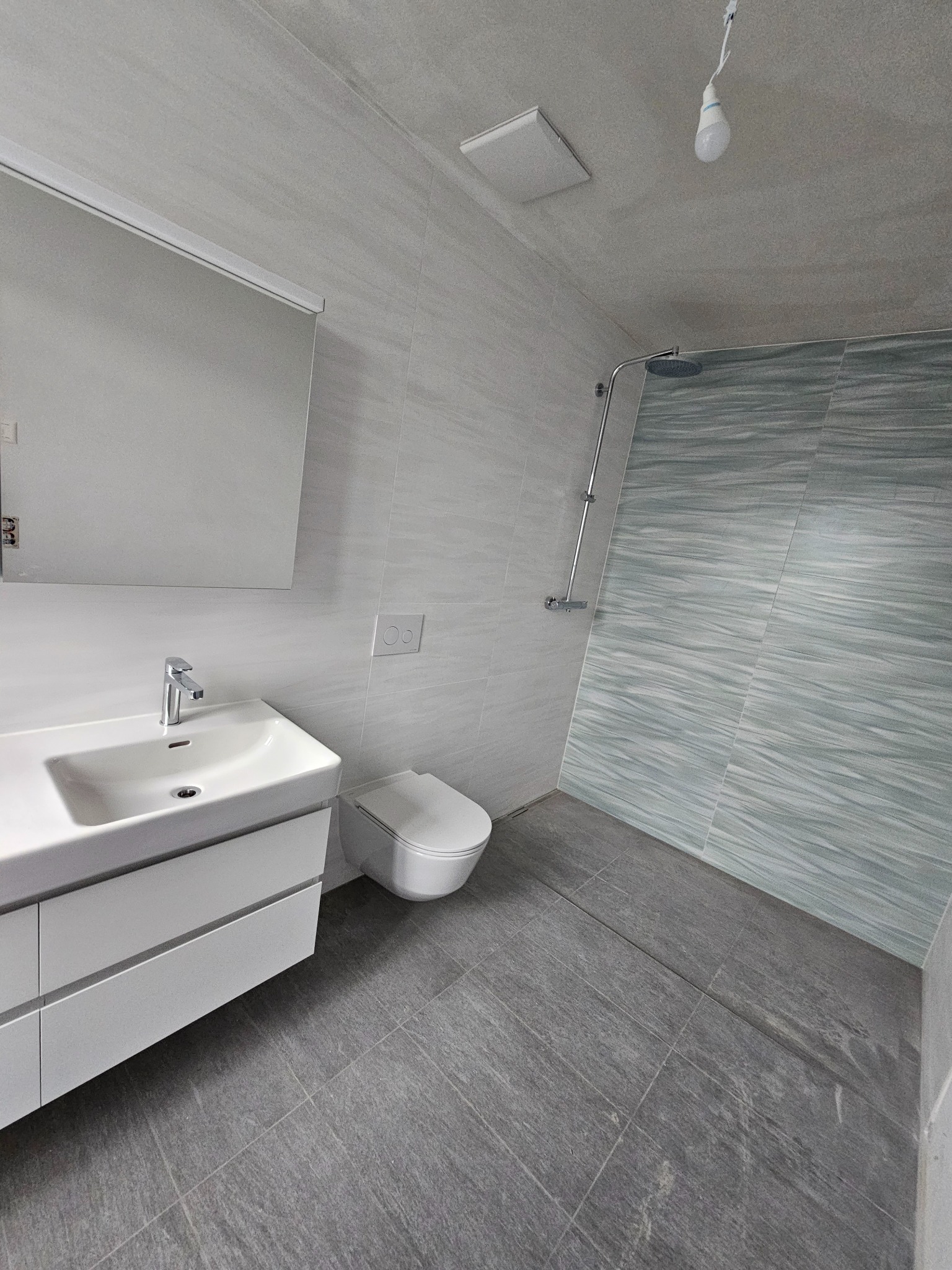 Nowoczesna łazienka w odcieniach szarości i bieli, z umywalką na szafce, toaletą i prysznicem z deszczownicą, wykończona płytkami imitującymi kamień i drewno.