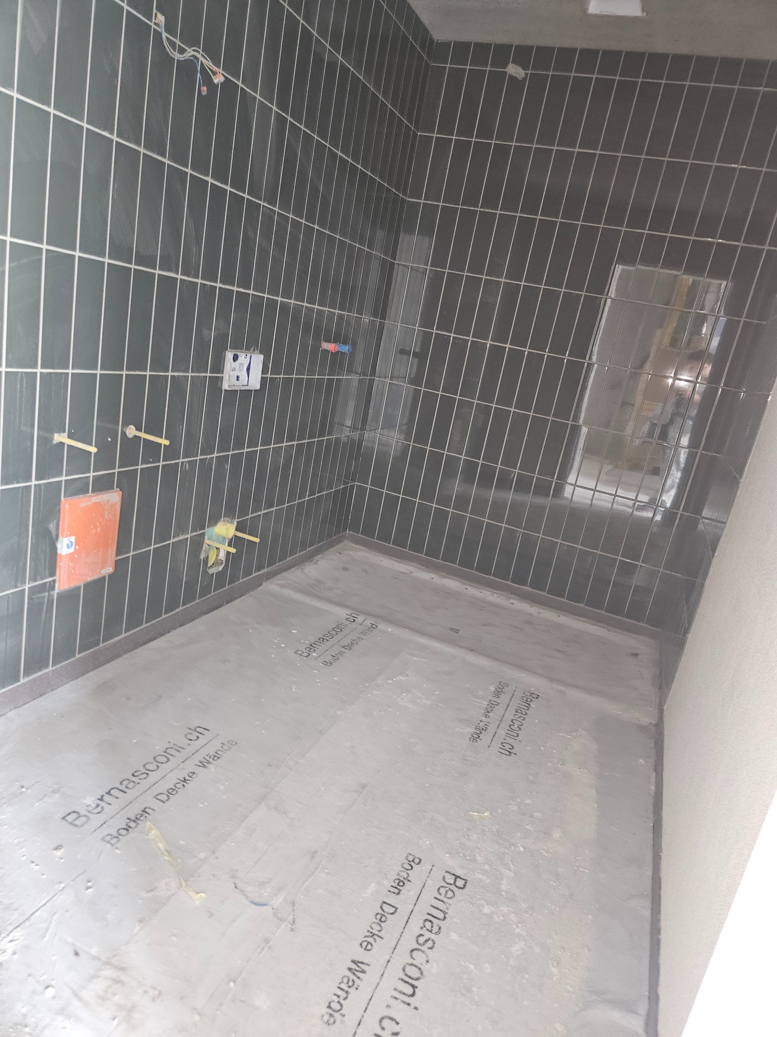 Łazienka w trakcie remontu: ciemne, błyszczące płytki ułożone na ścianach, przygotowane instalacje wodne i elektryczne, podłoga zabezpieczona folią ochronną z nadrukiem Bernasconi, widoczne...