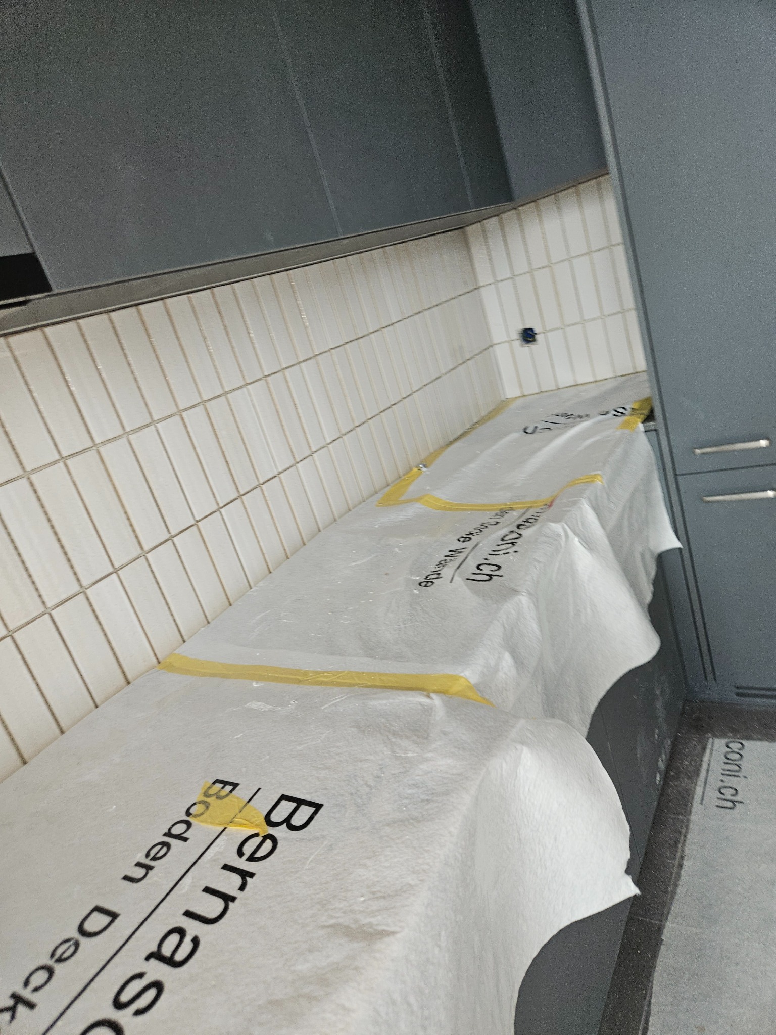 Przygotowanie do prac wykończeniowych: blat kuchenny zabezpieczony folią ochronną z nadrukiem, przyklejoną żółtą taśmą, widoczna ściana z białymi, wąskimi płytkami i szare szafki kuchenne.