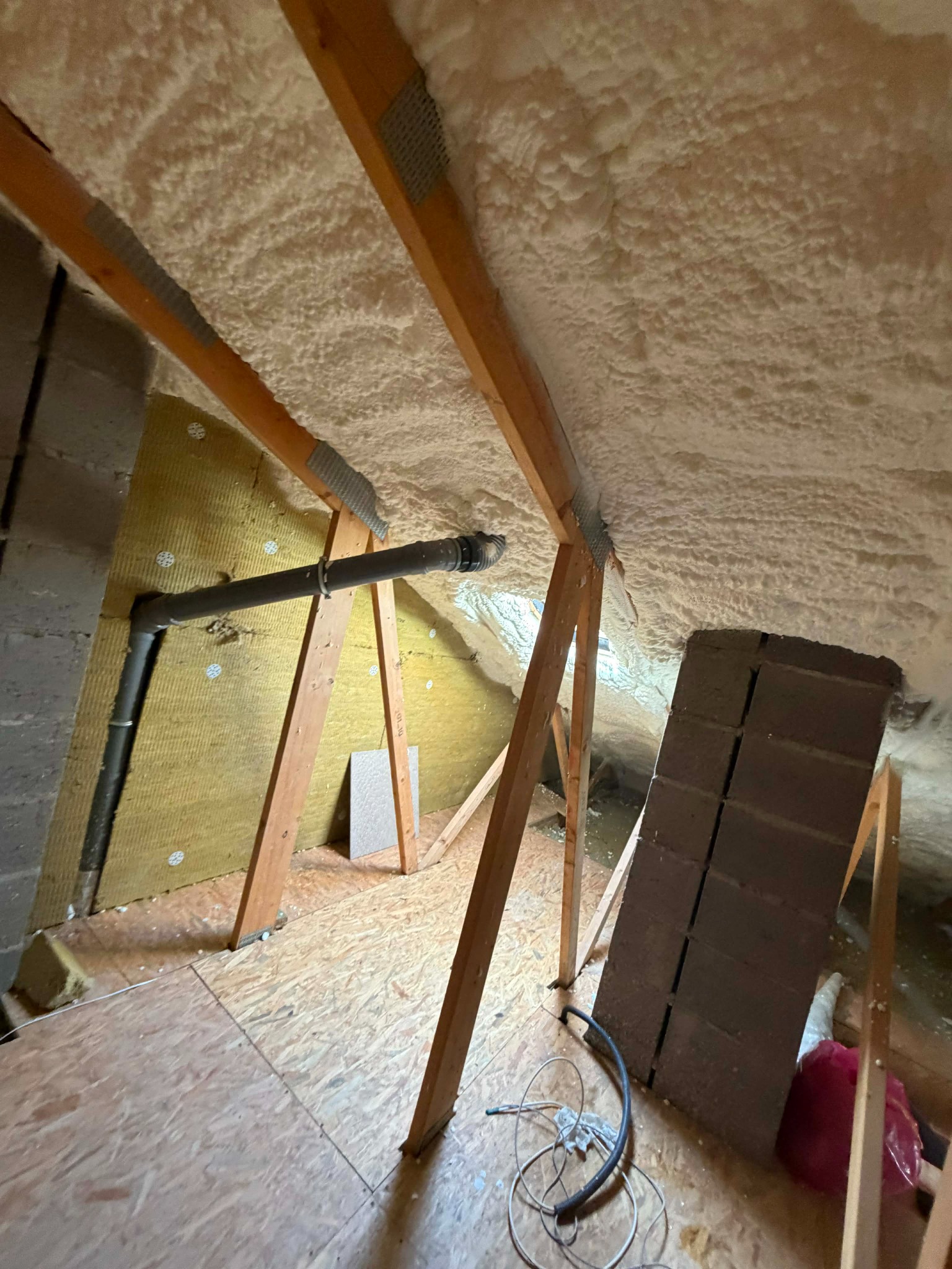 Poddasze po ociepleniu pianką PUR: widoczne drewniane krokwie, izolacja piankowa na skosach dachu, fragment instalacji wentylacyjnej i wełny mineralnej na ścianie szczytowej.