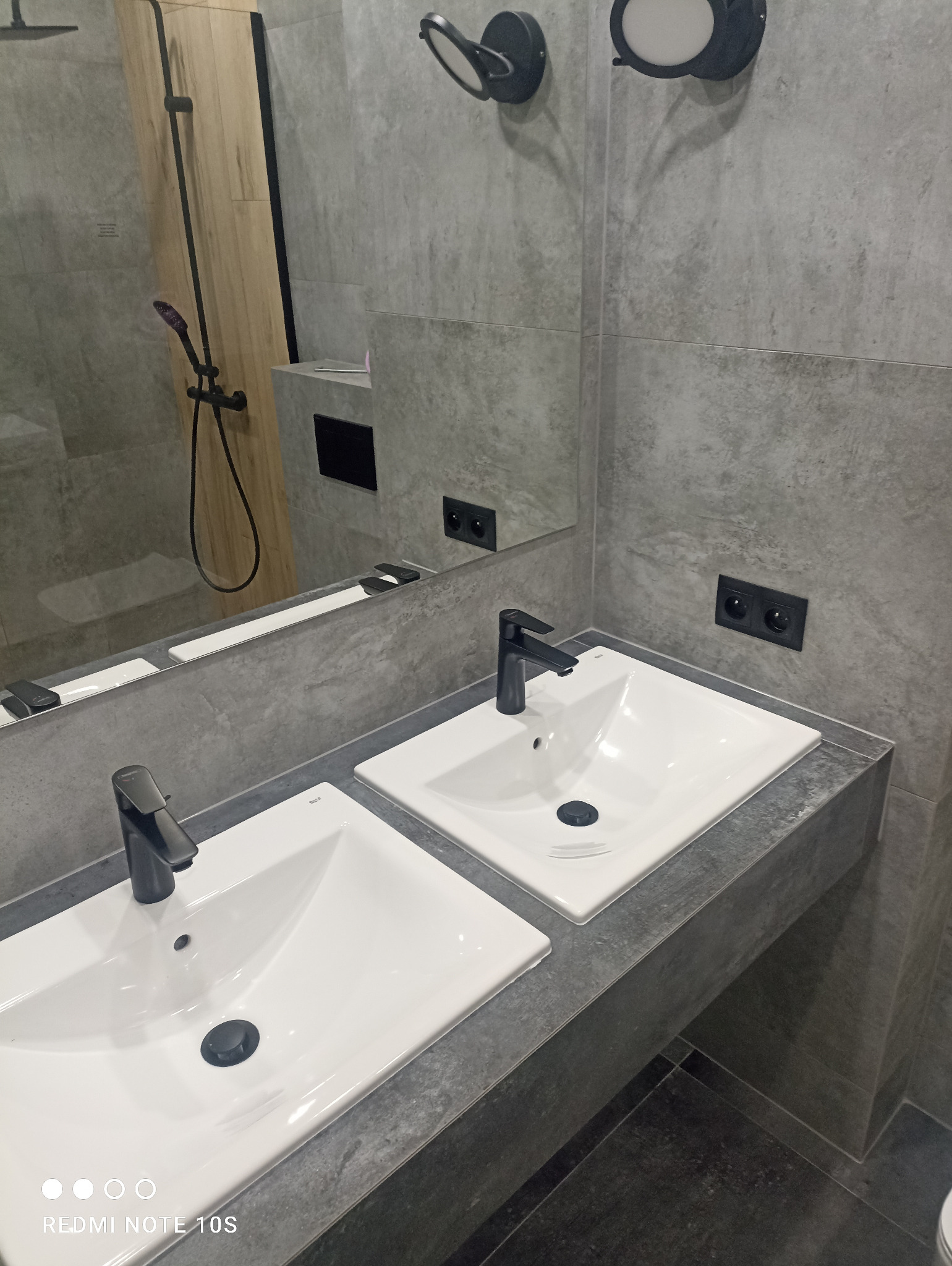 Nowoczesna łazienka z dwoma białymi umywalkami z czarnymi bateriami na blacie z szarego kamienia, szare płytki na ścianach, czarne gniazdka elektryczne i lustro odbijające prysznic.