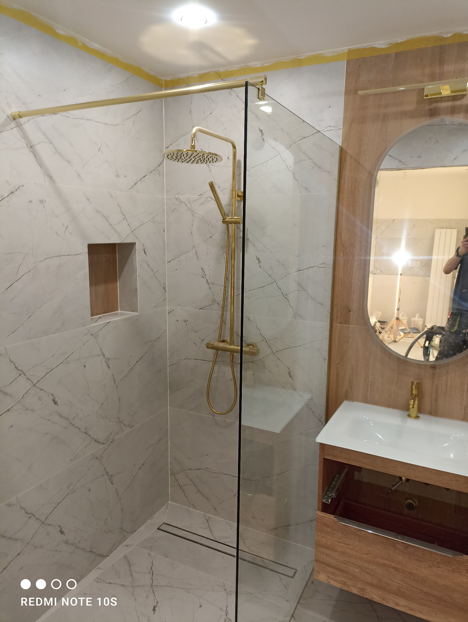 Nowoczesna łazienka z kabiną prysznicową typu walk-in, armaturą w kolorze złota i umywalką z szafką w odcieniach drewna, marmurowe płytki na ścianach z wnęką.