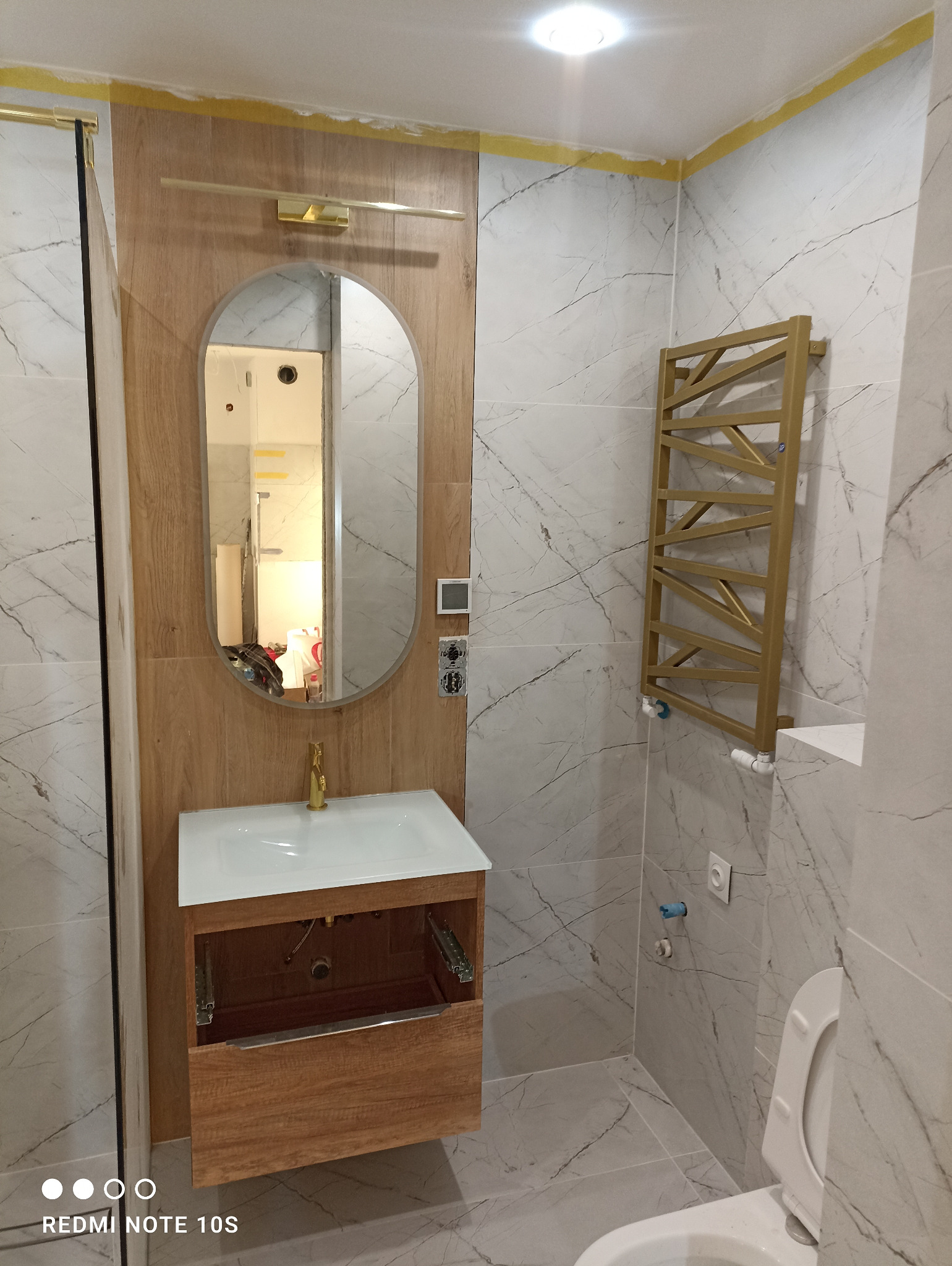 Nowoczesna łazienka z marmurowymi płytkami, drewnianą szafką pod umywalką z otwartą szufladą, owalnym lustrem z oświetleniem i złotym grzejnikiem drabinkowym.