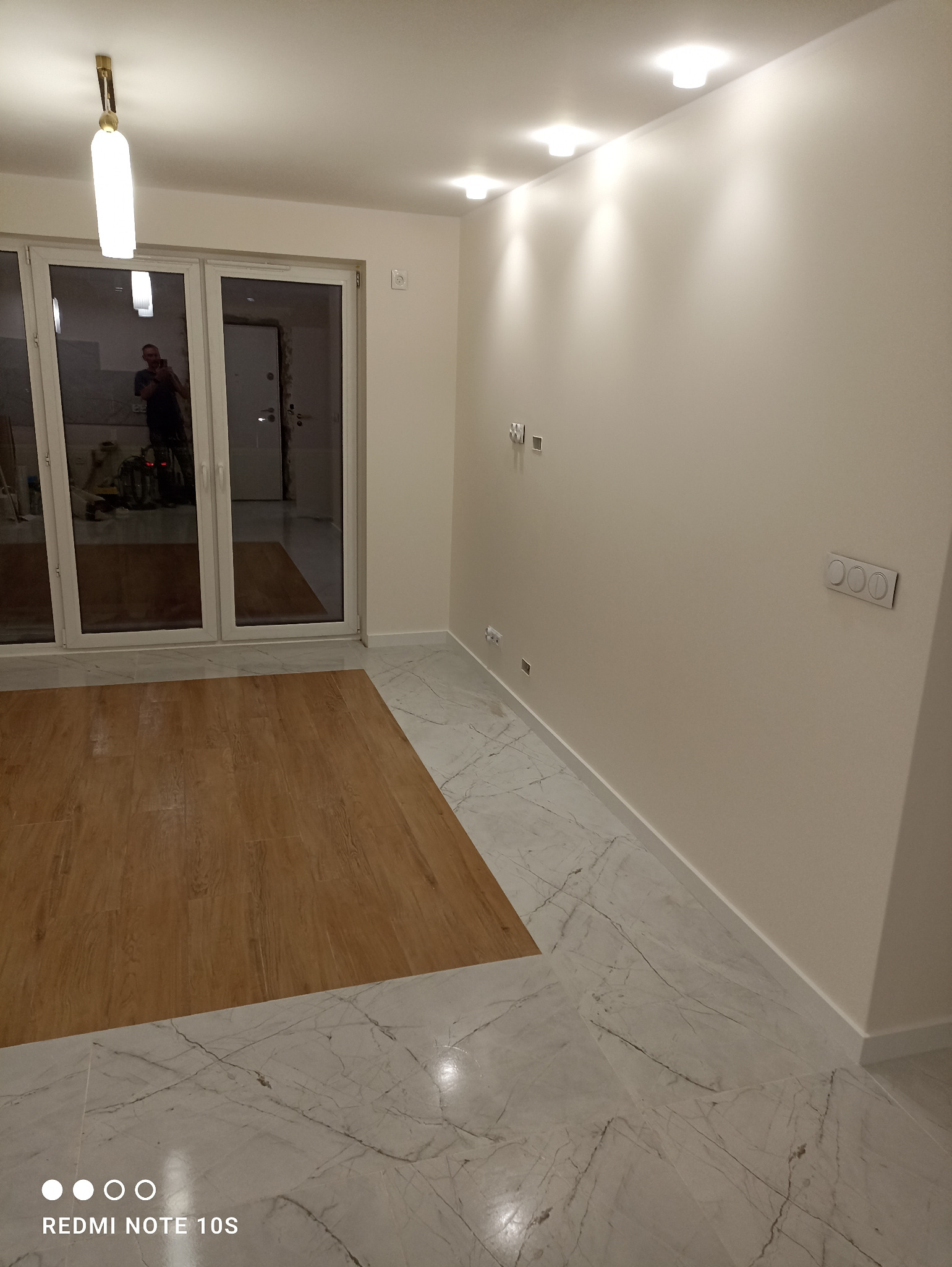 Jasne wnętrze po remoncie: połączenie paneli podłogowych imitujących drewno i marmurowej posadzki, białe ściany z nowoczesnym oświetleniem sufitowym i widocznymi gniazdkami elektrycznymi.