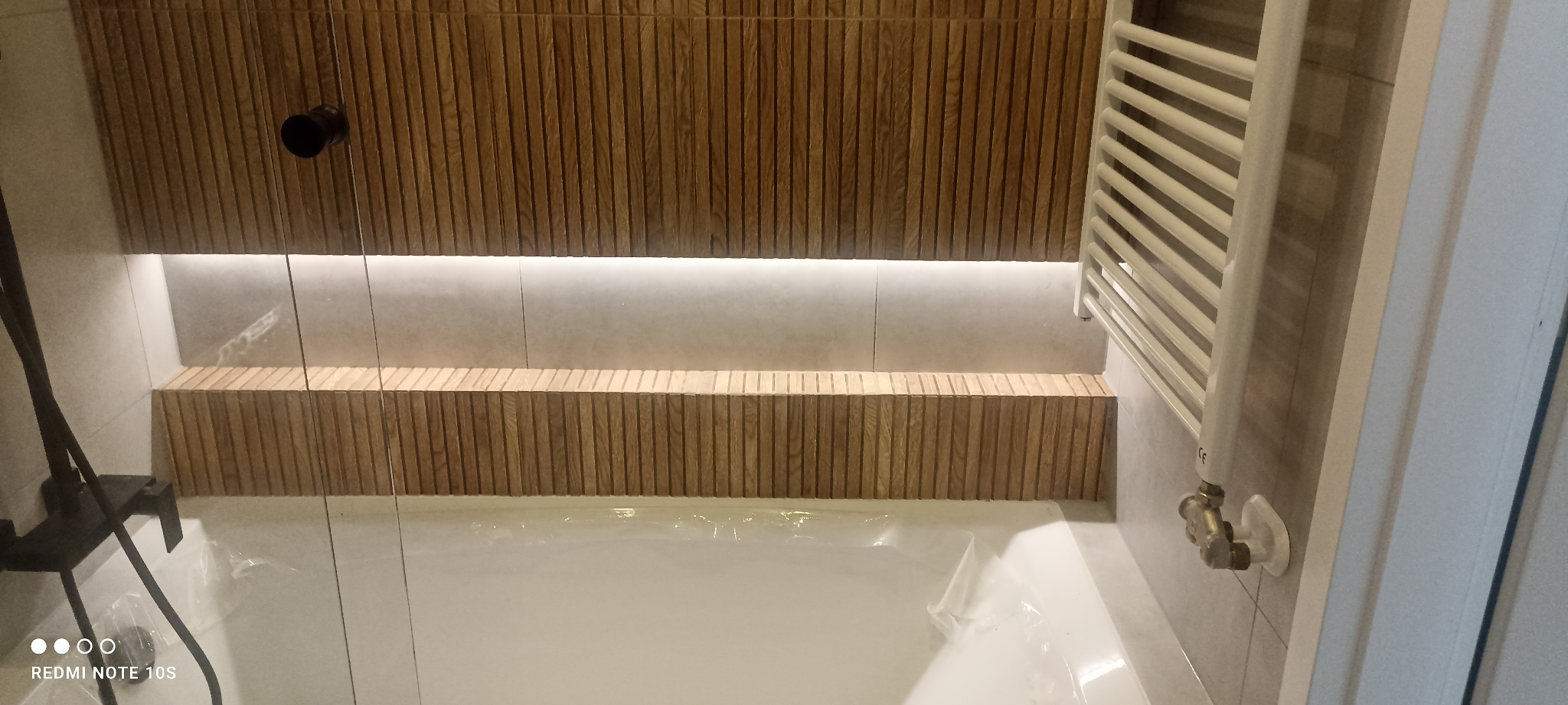 Wykończona łazienka z wanną zabezpieczoną folią, drewnianymi panelami na ścianie z podświetleniem LED i grzejnikiem drabinkowym.