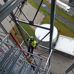 ALEX-BUD OLEKSANDR POPOVSKYI - Pracownik w zielonym kasku i jaskrawej kurtce zabezpieczony linami, wykonuje prace na konstrukcji stalowej wieży, widok z góry na parking, drogę i silos.
