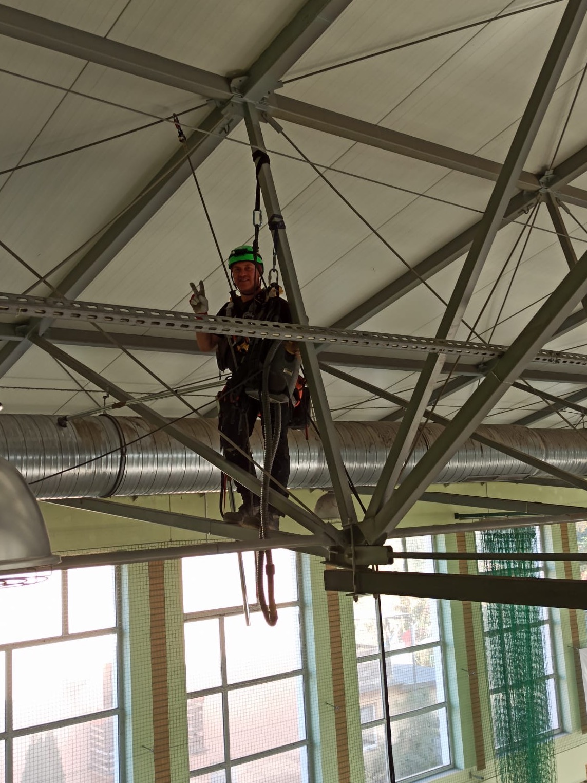 Pracownik w kasku i uprzęży, z uniesioną ręką w geście zwycięstwa, zawieszony na konstrukcji stalowej pod sufitem hali sportowej, obok widoczne rury wentylacyjne.