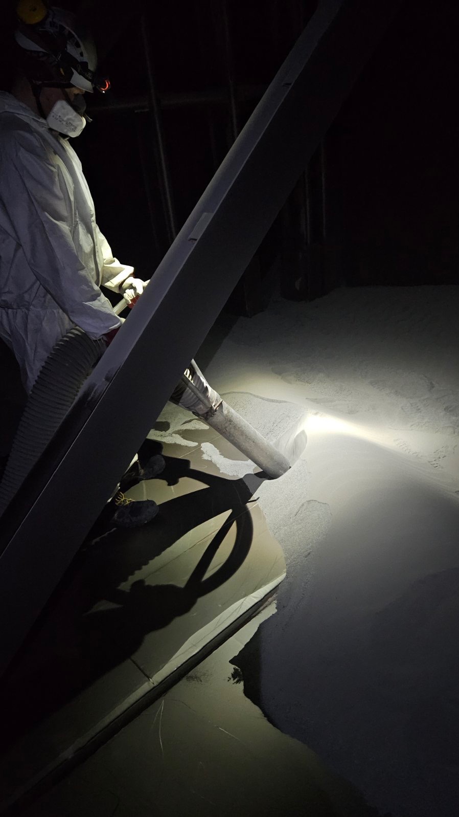 Pracownik w kombinezonie ochronnym usuwa biały pył z powierzchni za pomocą odkurzacza przemysłowego w słabo oświetlonym pomieszczeniu.