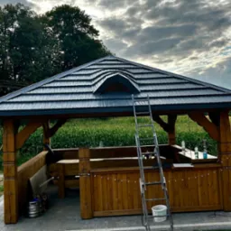 Zadaszona altana z drewnianą konstrukcją i czarnym dachem z blachodachówki, w Rozkopaczewie. Widoczna drabina i elementy dekoracyjne.