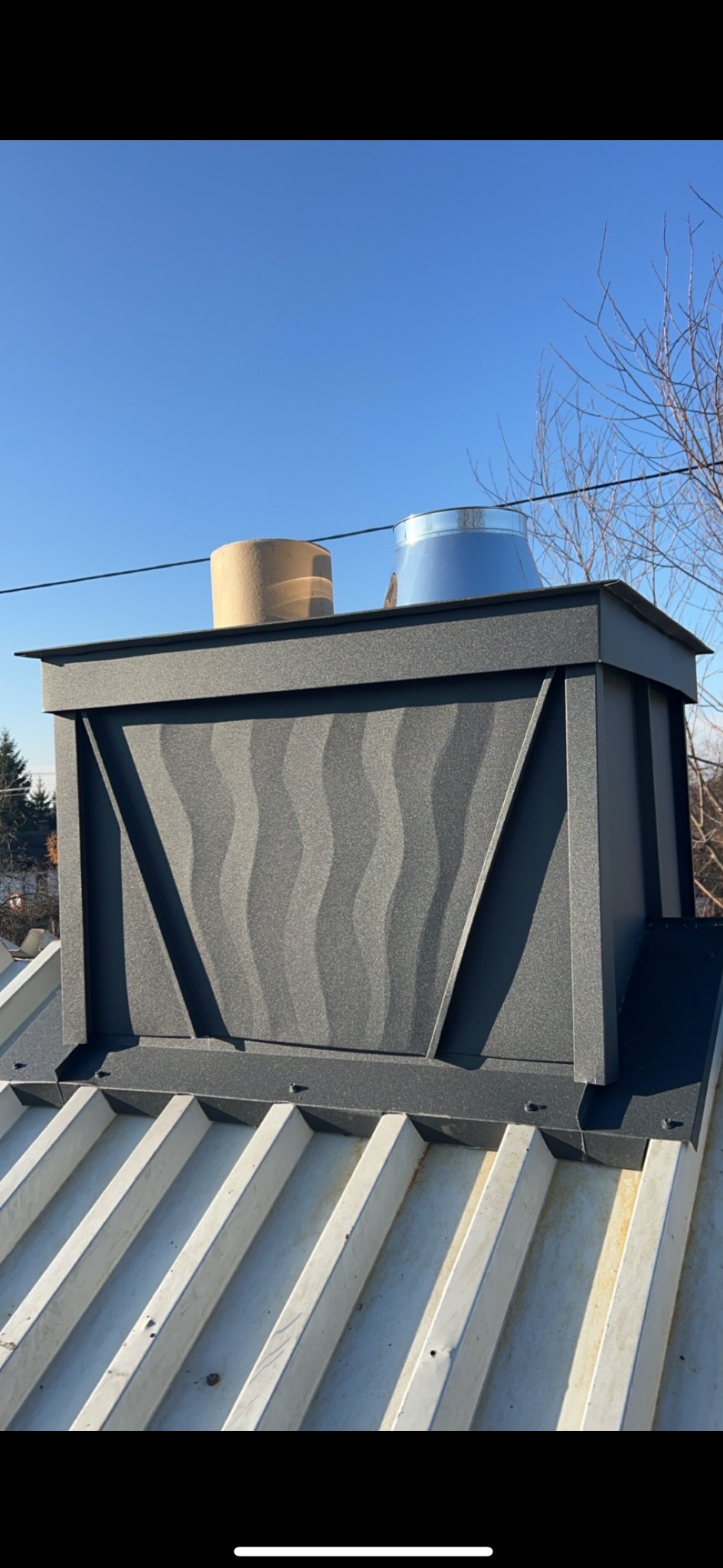 Szczegółowe ujęcie komina z czarną obudową o falistym wzorze, wystającego ponad białe, metalowe pokrycie dachowe, z widocznymi w tle elementami wentylacji i błękitnym niebem.