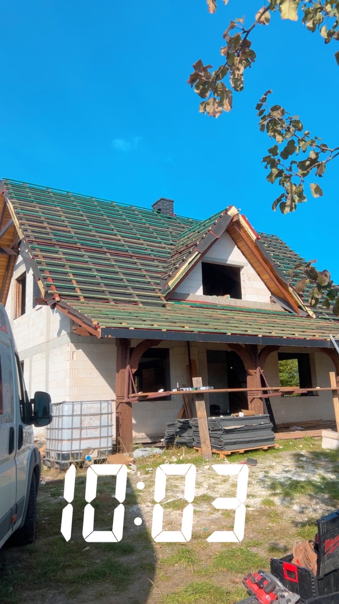 Dom w trakcie krycia dachu, widoczne łatowanie pod dachówkę, narzędzia pracy i fragment samochodu dostawczego. Ujęcie z dołu, błękitne niebo w tle.