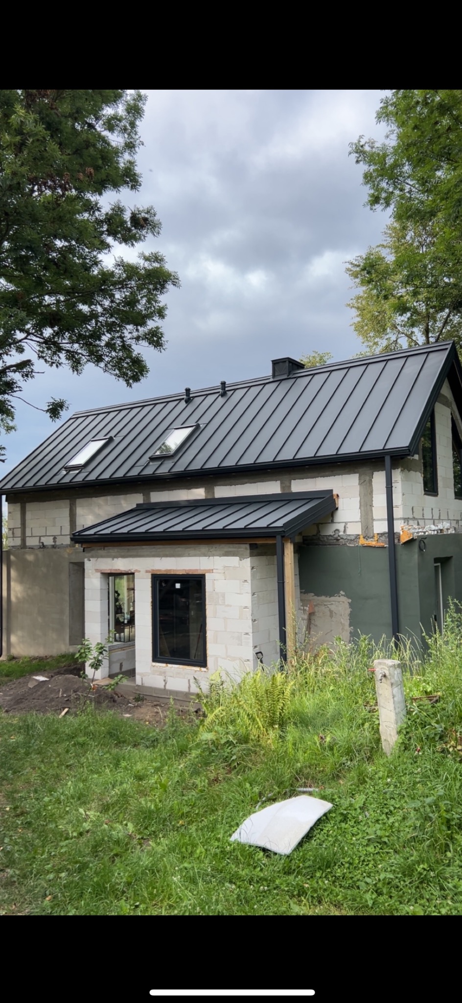 Dom w trakcie budowy z szarym dachem blaszanym i czarnymi rynnami. Widoczne okna dachowe i fragment elewacji z białego bloczka.