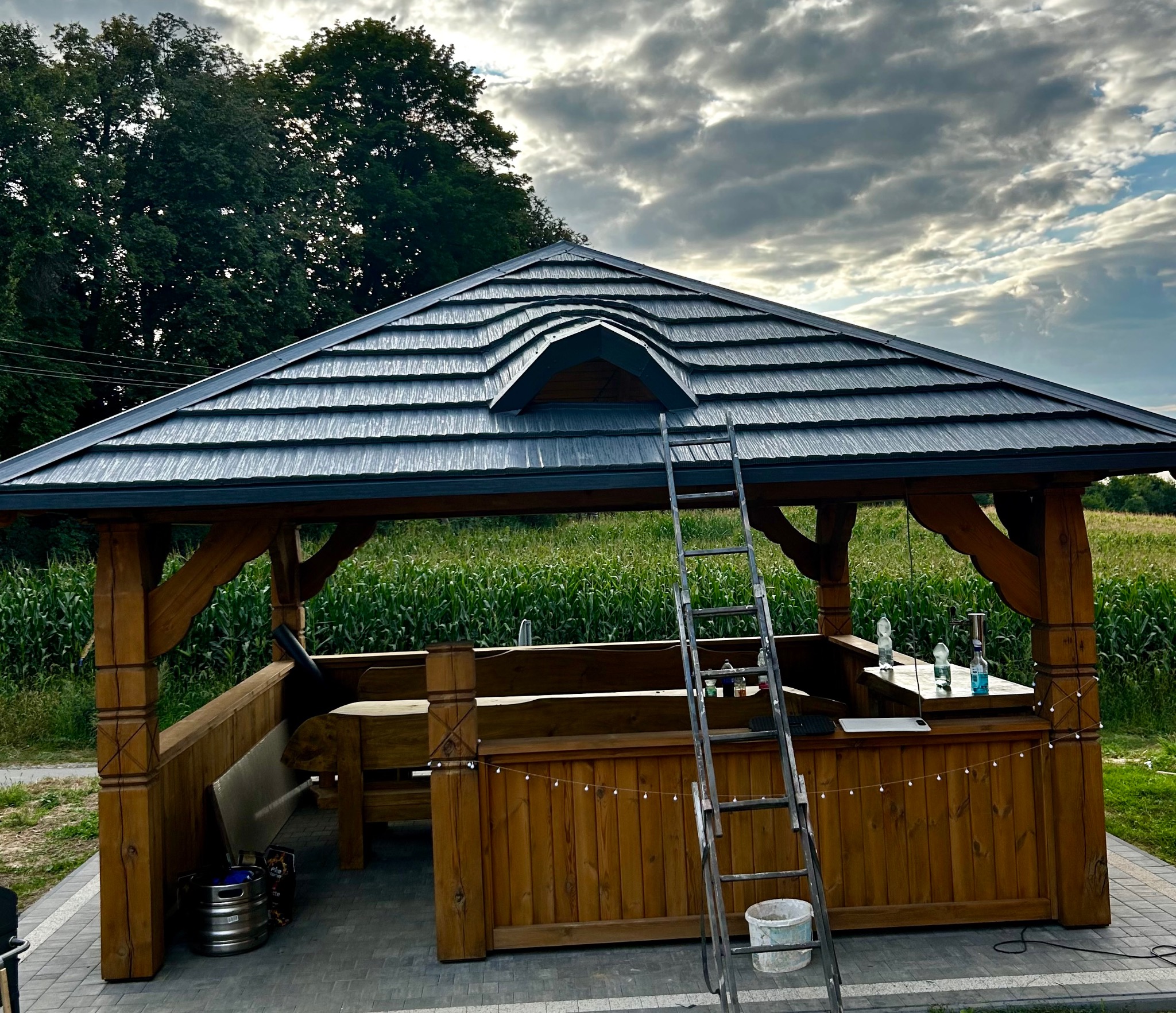 Zadaszona altana z drewnianą konstrukcją i czarnym dachem z blachodachówki, w Rozkopaczewie. Widoczna drabina i elementy dekoracyjne.