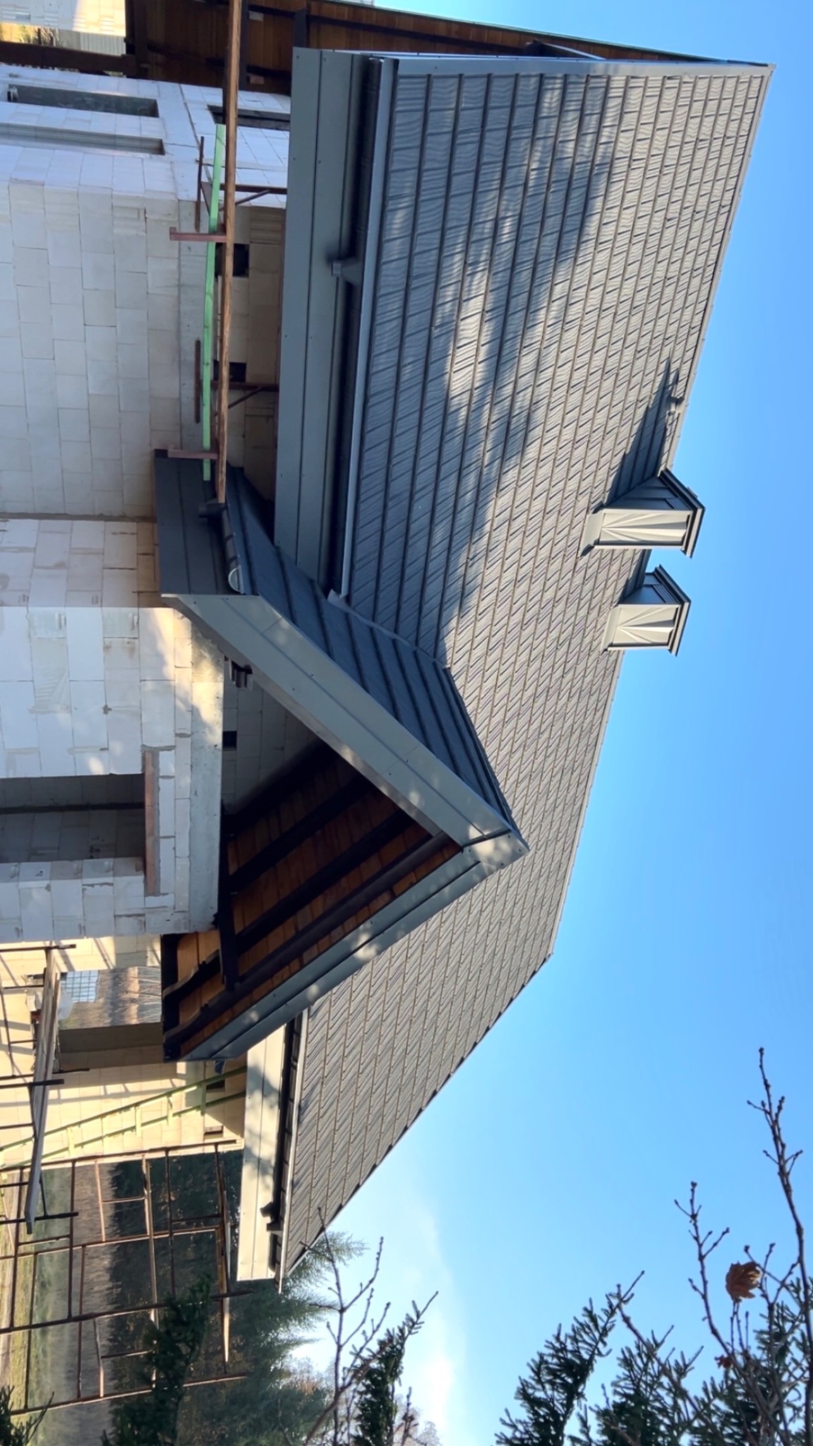 Szary dach z blachy na nowym domu w trakcie budowy, widoczne okna dachowe i fragment ściany z białego bloczka. Ujęcie z dołu, błękitne niebo w tle.