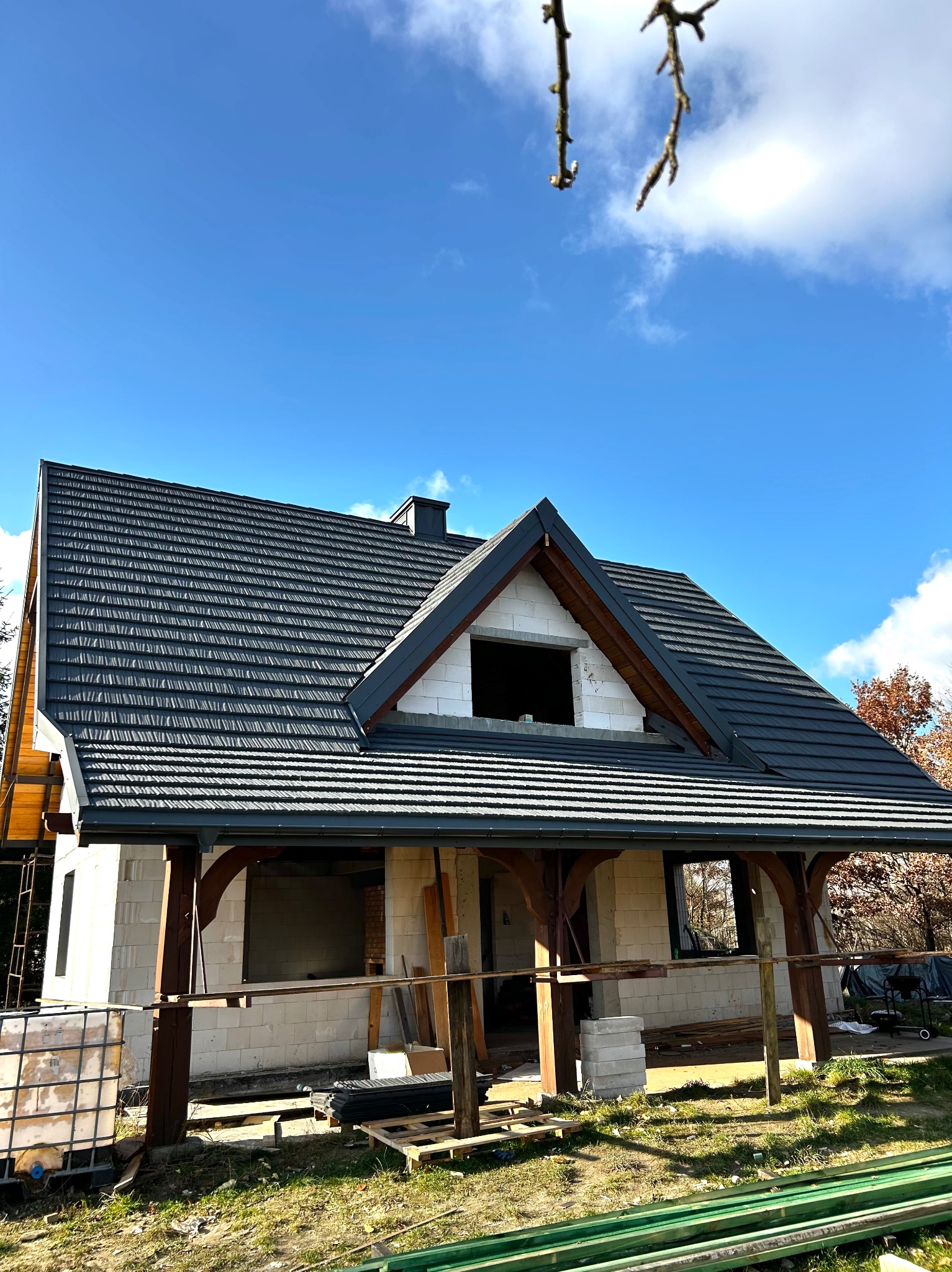 Dom w trakcie budowy z ciemnoszarym dachem z dachówki ceramicznej, widoczne elementy konstrukcyjne i rusztowania, błękitne niebo w tle.