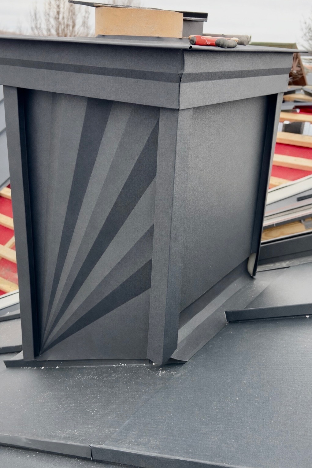 Szary komin wentylacyjny z dekoracyjnym wzorem na dachu z blachy na rąbek stojący. Widoczne narzędzia dekarskie i fragment więźby dachowej.