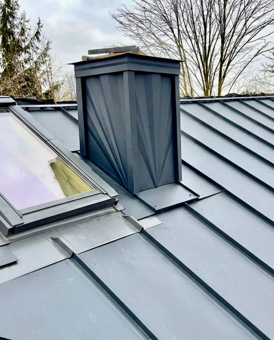 Szary dach z blachy na rąbek stojący z oknem dachowym i kominem z unikalnym wzorem, wykonany z blachy w tym samym kolorze. Detal obróbki blacharskiej.