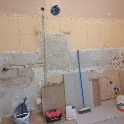 Kris-Bud - Fragment ściany w trakcie prac przygotowawczych do remontu: odsłonięte przewody elektryczne, częściowo usunięta stara farba, widoczne ślady po szpachlowaniu, narzędzia pracy i materiały budowlane...